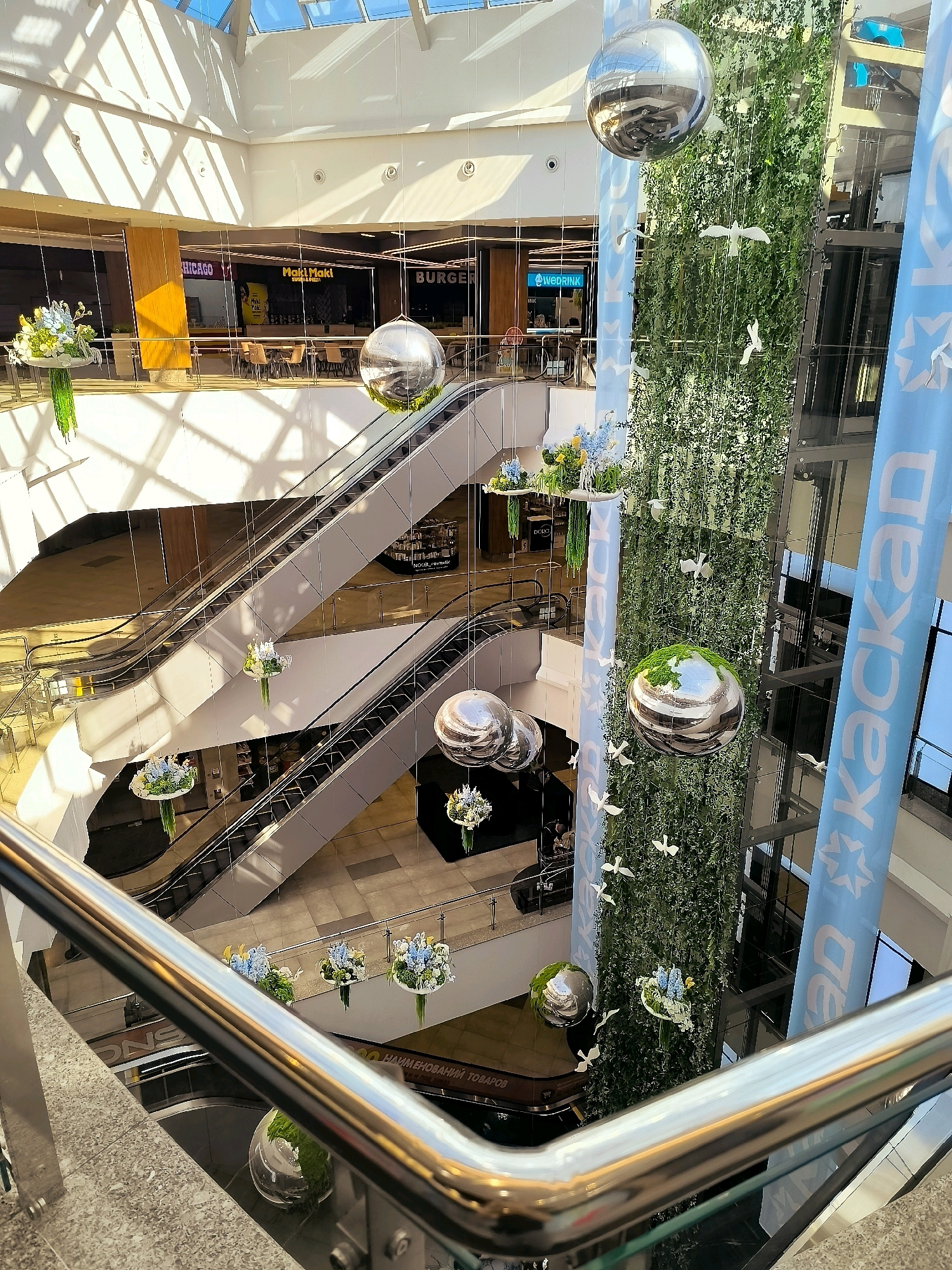 The image depicts the interior of a multi-level shopping mall. Escalators are visible, connecting different floors. Hanging decorations, including large mirrored spheres and floral arrangements, are suspended throughout the atrium. A vertical green wall with hanging white bird cutouts is a prominent feature. Large blue banners with white lettering, one of which reads "kackan", are displayed vertically along an elevator shaft. The architecture features a large glass ceiling with sunlight streaming through, casting shadows. Shops with signage such as "CHICAGO", "Maki Maki", and "BURGER" are visible on the upper levels. A stainless steel railing is in the foreground. The scene appears to be a public commercial space within Qarağandı city administration, Kazakhstan.