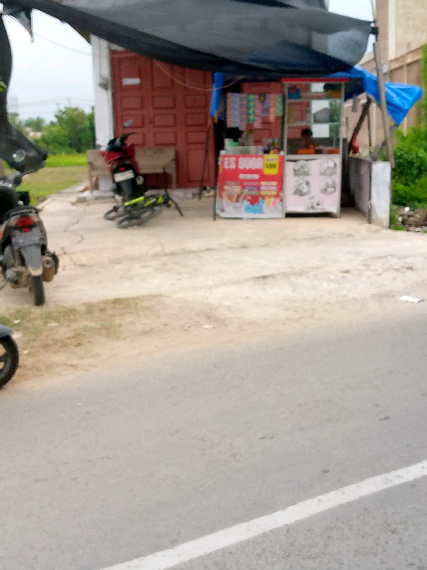 The image shows a small food stall on the side of a road in Mesjid, Indonesia. The stall appears to be selling iced beverages, as indicated by the signage that reads "ES BOGOR" and depicts images of colorful drinks. A person is partially visible behind the counter of the stall, presumably the vendor. In front of the stall, there is a motorcycle parked, and to the left of the stall, another motorcycle is visible from the rear. A bicycle is also present, leaning on the ground near the stall. The ground in front of the stall is paved with concrete, and the road in the foreground is asphalt with a white line marking. Lush green vegetation is visible in the background, suggesting a semi-urban or rural setting. The stall is covered by a dark canopy. The overall scene depicts a typical street-side vendor operation in Indonesia.