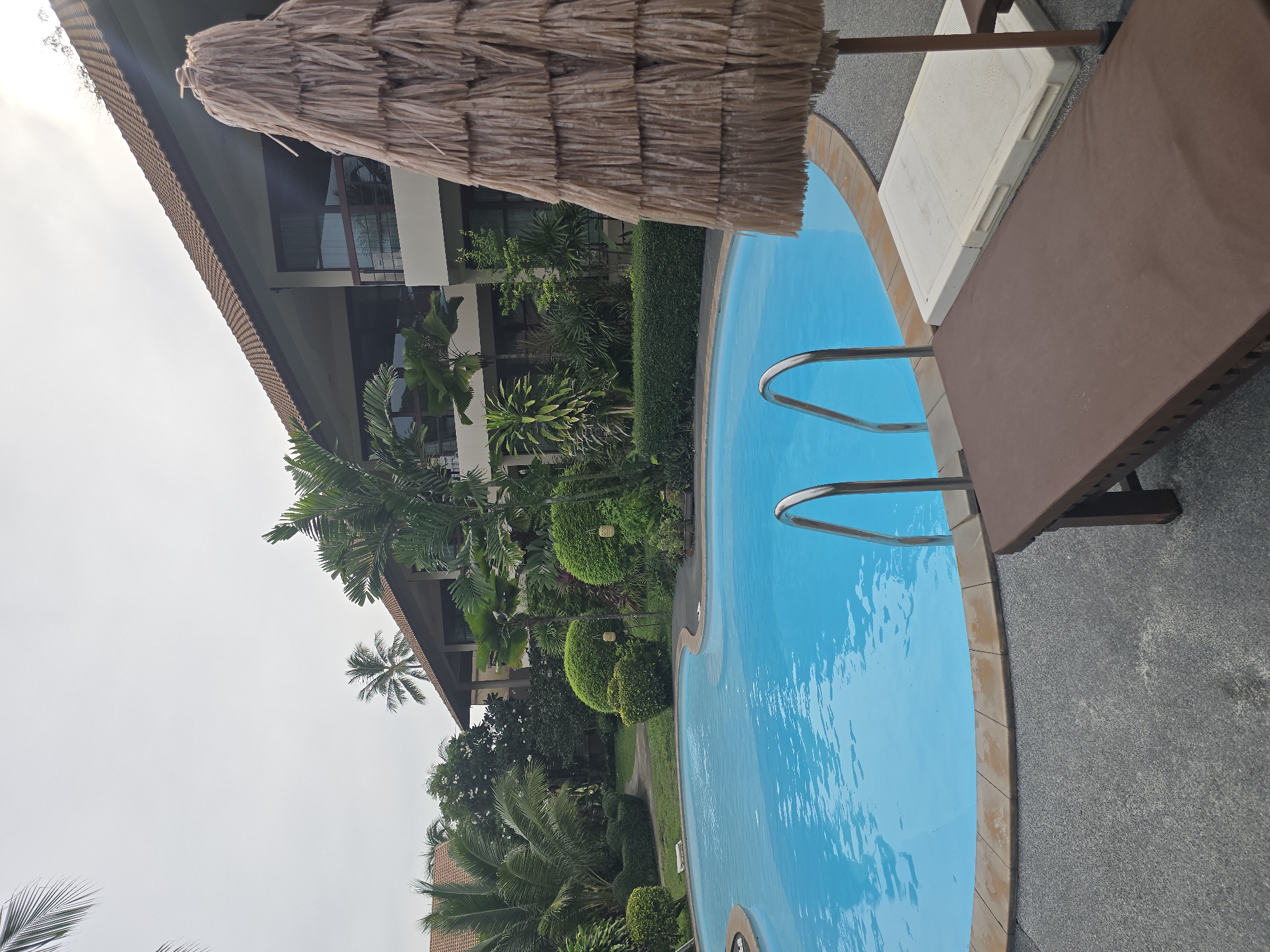 An outdoor shot shows a swimming pool with bright blue water and a brown tiled edge. A set of silver pool stairs leads into the water from the left side of the frame. To the right of the pool, a brown lounge chair sits on a gray, speckled concrete surface. Behind the lounge chair, a straw umbrella is visible. In the background, a multi-story building with a brown tiled roof and balconies is surrounded by lush green vegetation, including palm trees and manicured bushes. The sky is overcast and gray, suggesting a cloudy day. The scene appears to be a resort or hotel.