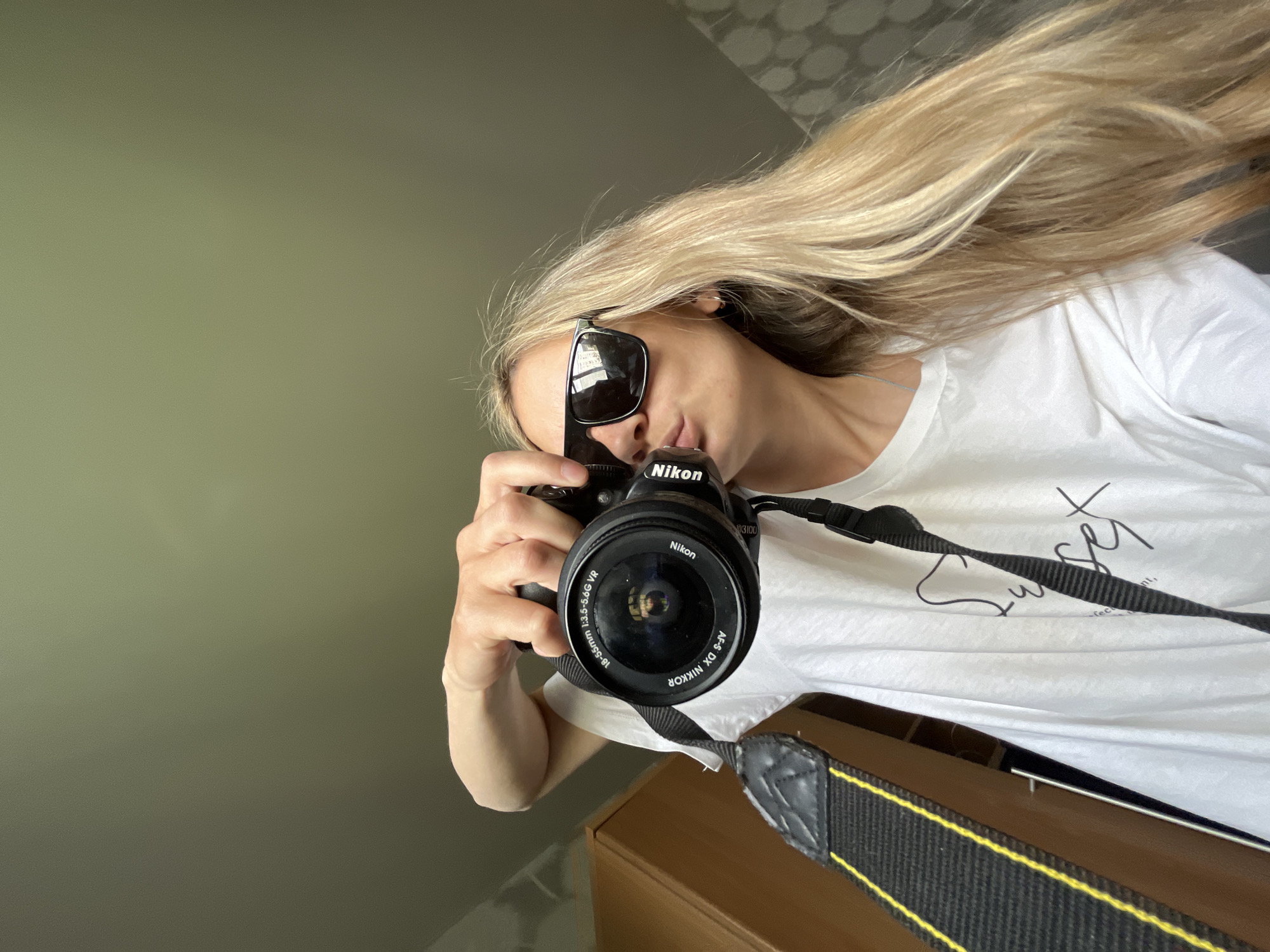 A woman with long, blonde hair and sunglasses is holding a Nikon camera in front of her. The camera has a black strap with a yellow stripe. She is wearing a white t-shirt and is partially visible from the chest up. The background includes a light green wall and a brown cabinet. It's unclear if the location is Chaikovsky, Russia, as there are no immediately identifiable landmarks or signs.