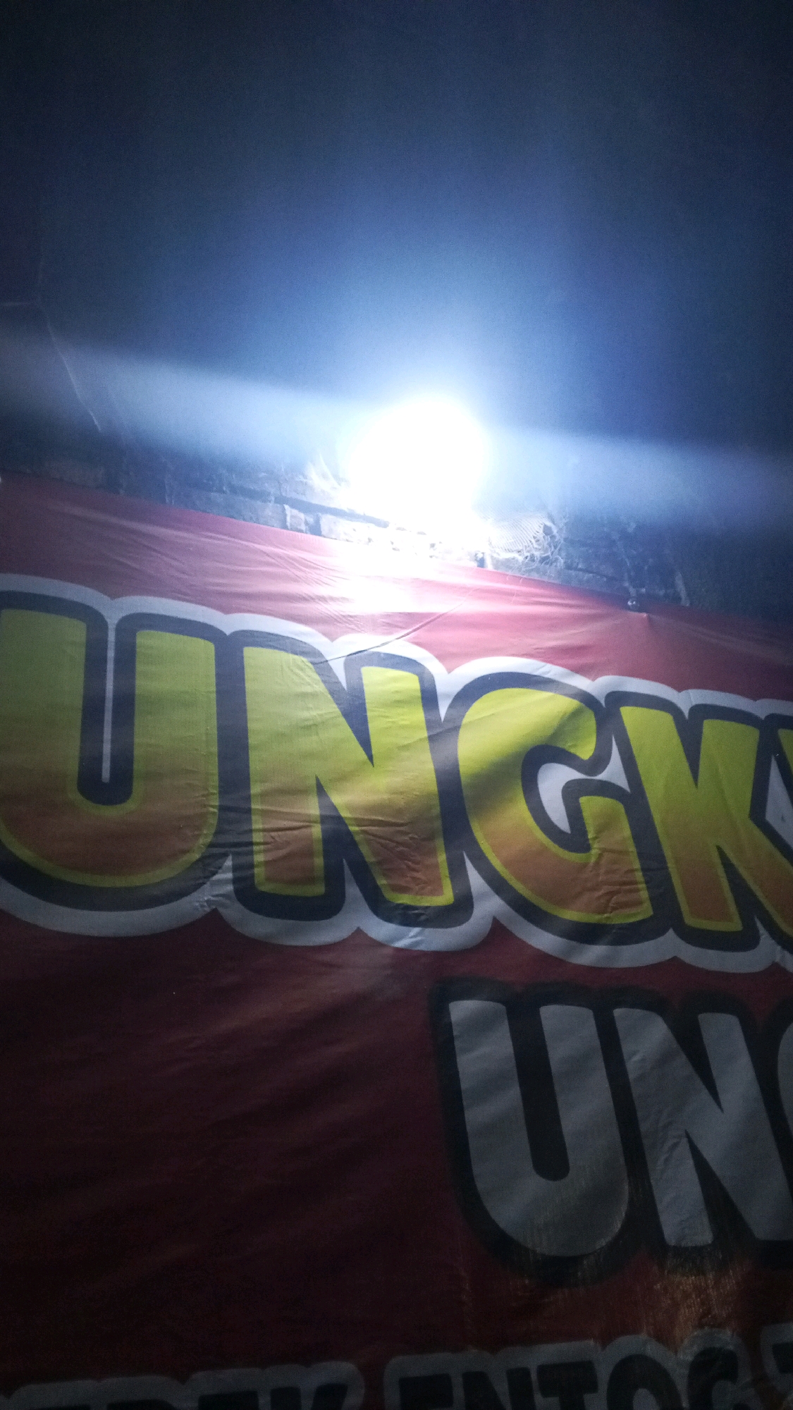 A close-up, eye-level, dimly lit shot shows a red banner with large yellow, black, and white lettering. The banner is stretched taut and appears to be outdoors, possibly at night given the bright overhead light source casting a glare. The text "UNGK" is clearly visible in the upper portion of the banner, with the bottom of the banner showing the beginning of the word "UN" in large gray letters. The banner is likely advertising an event or business in Kalinyamatan, Indonesia.