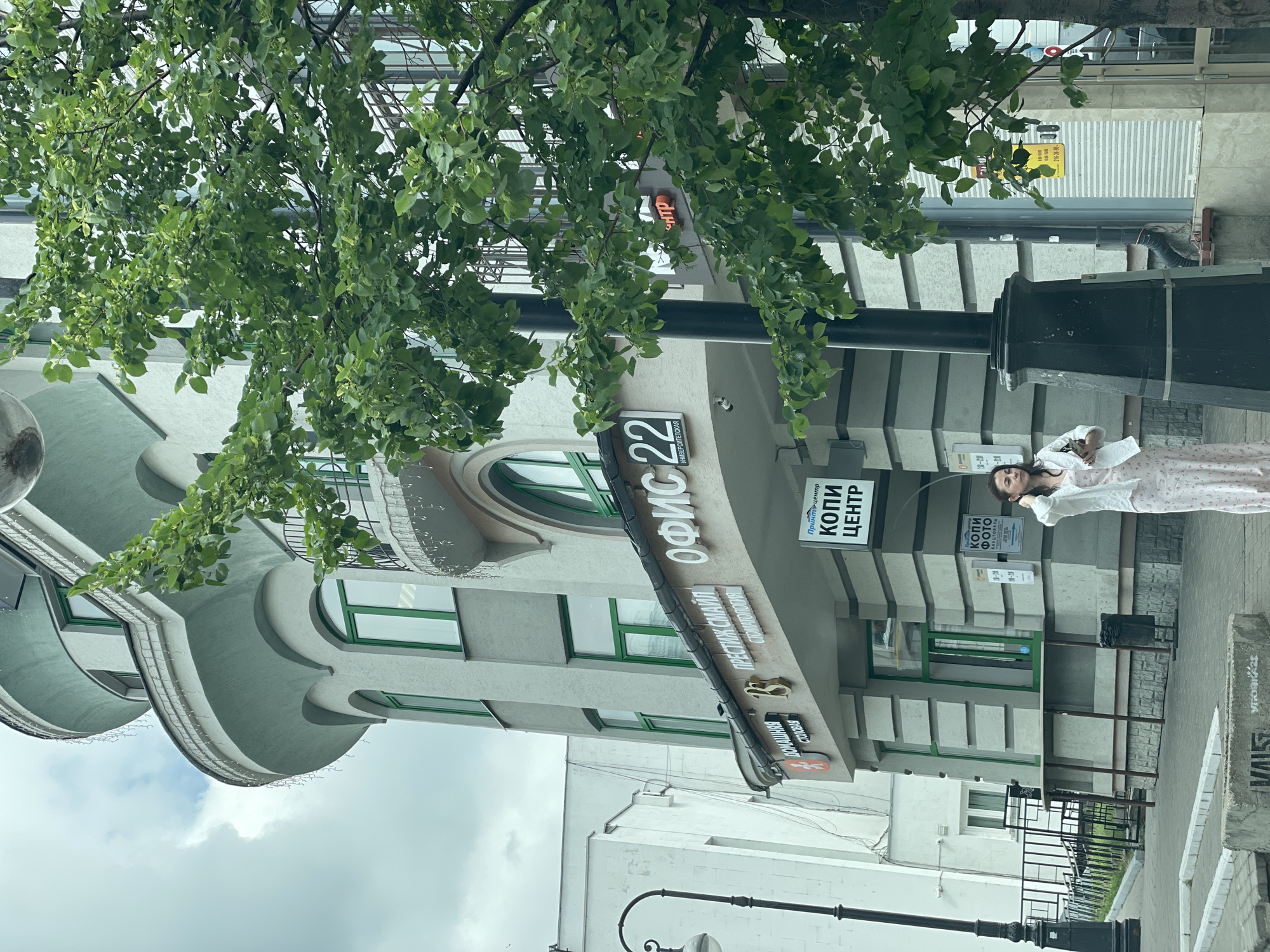 An exterior, low-angle, medium shot captures a woman in a light-colored, floral print dress and white cardigan walking on the sidewalk in front of a multi-story building with light gray and mint green accents. The building has arched windows with green frames and decorative balconies. Signs on the building include "ОФИС 22" (Office 22), "ПРЕСТИЖ СМАЙЛ" (Prestige Smile), "СТОМАТОЛОГИЯ" (Dentistry), and "КОПИ ЦЕНТР" (Copy Center). A large tree with green leaves is in the foreground, partially obscuring the building. A black streetlamp is also visible. The background shows a cloudy sky and other buildings. The scene is located in Kazan, Russia.