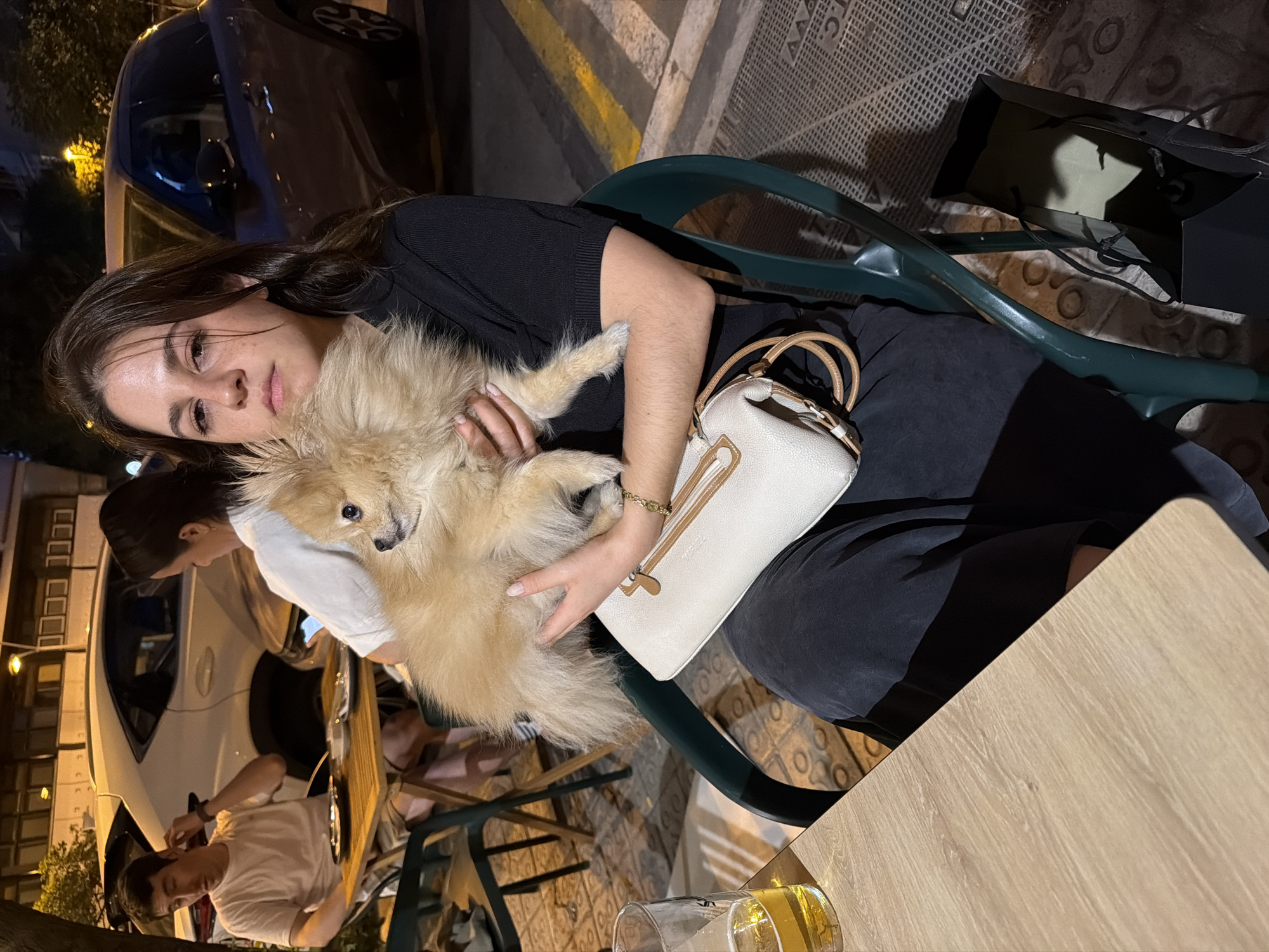 A young woman with dark brown hair is sitting in a dark green chair, holding a small, fluffy, light brown Pomeranian dog in her arms. She is wearing a black short-sleeved shirt and a dark skirt. In her arms, she holds a white clutch purse with light brown trim. A glass of amber liquid, likely beer, sits on a light brown wooden table to her left. In the background, a man wearing a beige t-shirt and shorts is seated at a similar table, looking to his left and touching his ear. Behind him, a white SUV is parked. Further in the background, a dark blue SUV is also parked. The scene appears to be an outdoor seating area of a restaurant or cafe, with a tiled ground and a street visible. The presence of parked cars and buildings suggests an urban environment. The lighting indicates it is nighttime or dusk. The image was captured in Barcelona, Spain, as evidenced by the car models and architectural style consistent with the city.