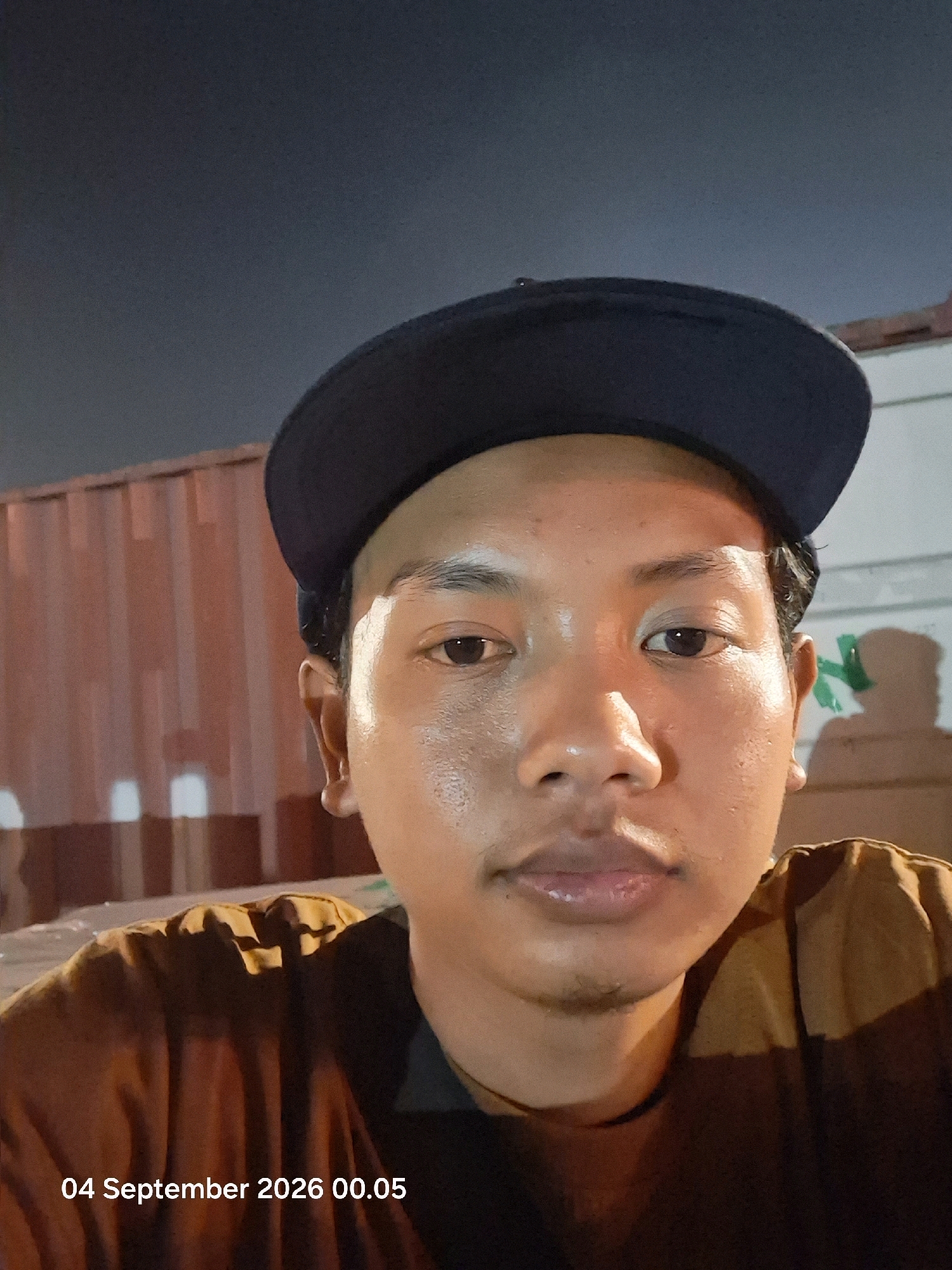 A close-up, waist-up selfie taken at night shows a person wearing a dark blue baseball cap and a brown t-shirt. The person has dark hair and appears to be sweating. The background consists of a corrugated metal wall and a dark sky. The image is watermarked with "04 September 2026 00.05". The location is stated to be Guntung River, Indonesia.