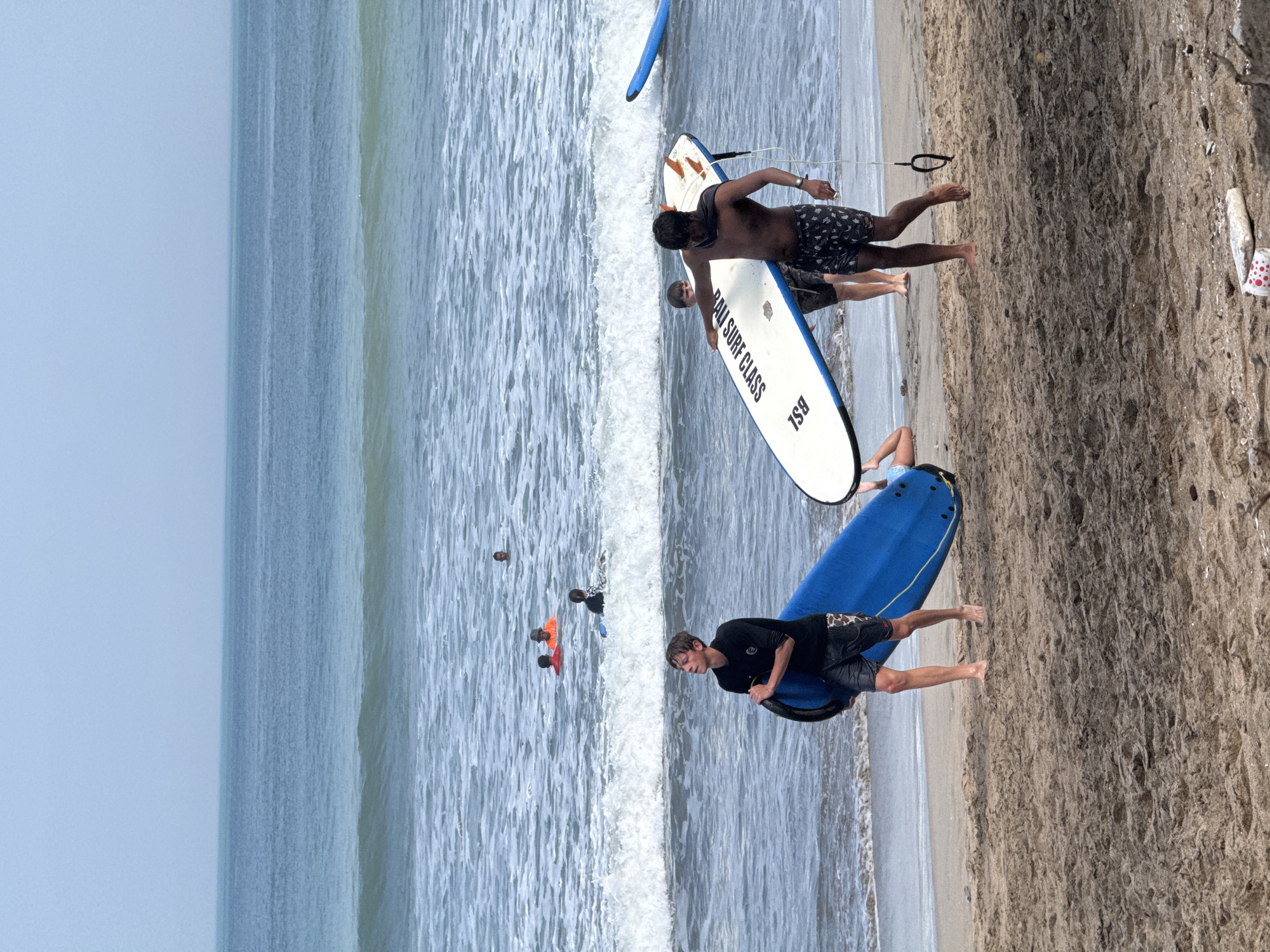 The image depicts a beach scene in Kuta, Indonesia, likely during a surf lesson. The ocean is calm with small waves breaking near the shore. Several people are in the water, some with surfboards, while others are simply swimming or wading. On the beach, two young men are prominent. One, wearing a black long-sleeved shirt and black and white patterned shorts, carries a blue surfboard and a smaller blue board under his arm, looking up and to his left. The other, shirtless and wearing patterned shorts, carries a white surfboard with blue trim that reads "BALI SURF CLASS" and "BSL". Another person, possibly a child, is partially visible lying on the sand near the water's edge behind him. In the background, several other individuals are visible in the ocean, some in orange or red tops, indicating they might also be part of a group or lesson. The sand on the beach is light brown and appears somewhat damp near the water. The sky is overcast and a pale blue, suggesting a diffused light. A few pieces of debris are scattered on the sand in the foreground.