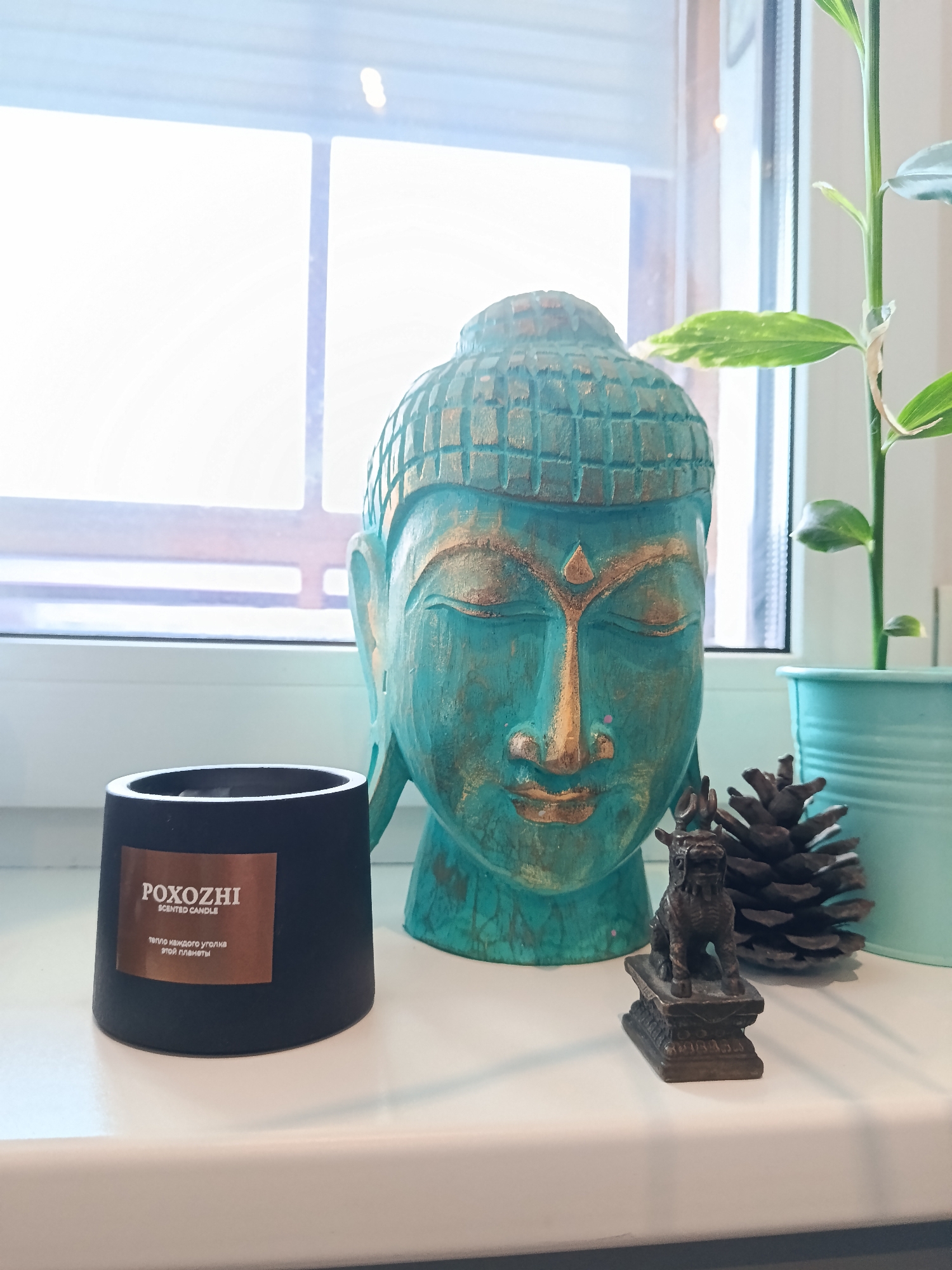 The image presents a static, indoor still life arrangement on a white windowsill. The central subject is a large, decorative Buddha head statue, painted in a distressed teal-green color with gold accents on its facial features and hair.
To the left of the Buddha head is a black, cylindrical scented candle, likely made from a cement-like material. It bears a copper-colored label with text: "POXOZHI," "SCENTED CANDLE," and in Cyrillic, "ТЕПЛО КАЖДОГО УГОЛКА ЭТОЙ ПЛАНЕТЫ," which translates to "THE WARMTH OF EVERY CORNER OF THIS PLANET."
Further to the right, in front of the Buddha head, is a small, dark, metallic figurine resembling a Foo dog or a mythical creature with horns, sitting on a base. Adjacent to it is a natural, dark brown pine cone. On the far right, a potted plant with green and variegated leaves stands in a ribbed, light blue pot, partially visible.
The background is dominated by a large window, through which bright, diffused white light enters, indicating it is daytime. The soft light suggests either an overcast sky or indirect sunlight. No specific activity is taking place; the scene is purely a decorative display. There are no people visible. The arrangement of objects conveys a sense of calm and aesthetic appeal, likely intended for home decor.