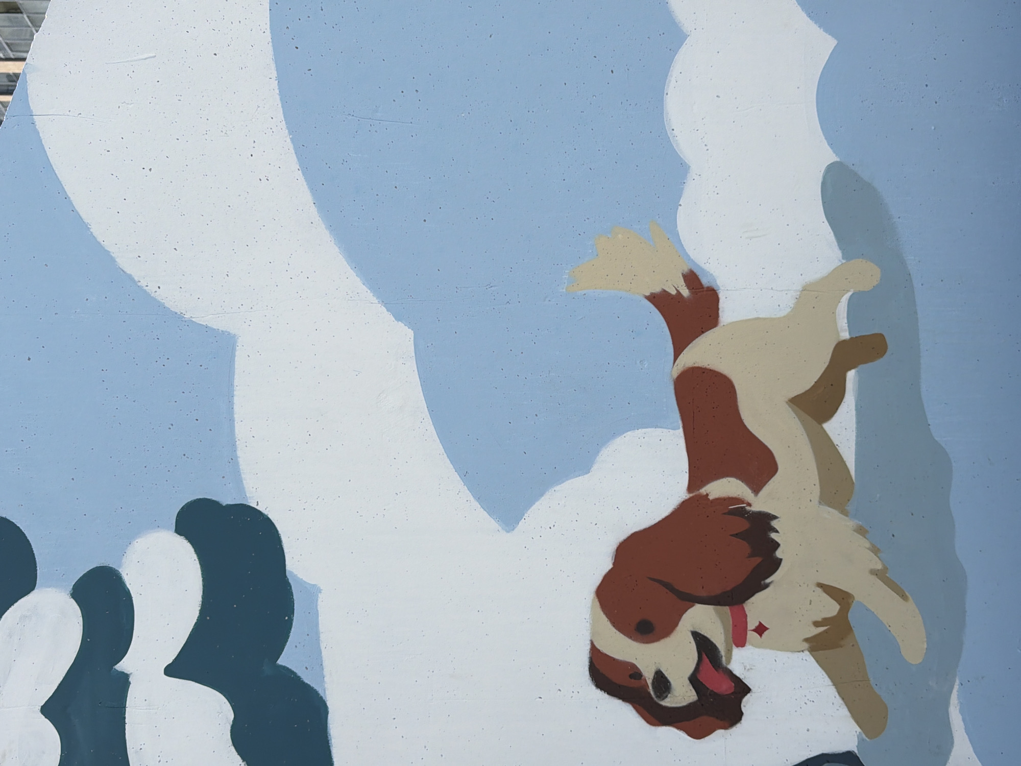 The image shows a section of a mural painted on a wall, depicting a cartoon-style dog running through a snowy, abstract landscape. The dog is brown and white with floppy ears and a pink collar. The background consists of large, puffy white shapes that appear to be snowdrifts or clouds, set against a light blue sky. A dark green and white shape in the upper left corner might represent a snow-covered tree or shrub. The mural is rendered in a flat, stylized manner. There is no discernible information within the image to indicate its specific location in Yekaterinburg, Russia.