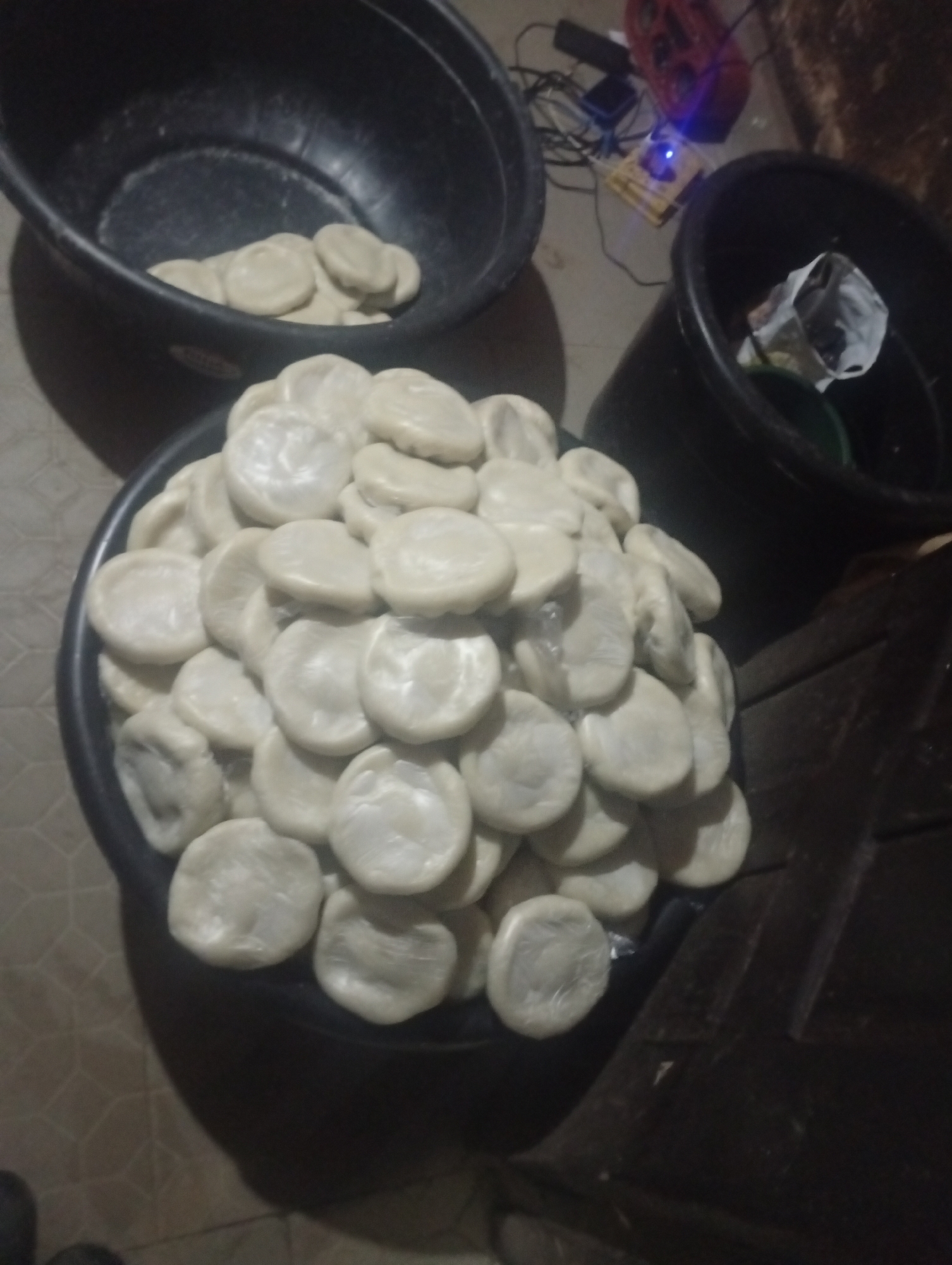 In Bukuru, Nigeria, a close-up shot captures a large, dark basin overflowing with numerous round, flat, pale objects, possibly food items like dumplings or dough balls. These items are stacked densely, filling the basin to its brim. To the left, another dark basin is partially visible, also containing a smaller pile of these pale objects. In the background, a collection of electronic devices, including what appears to be a power strip with a blue light, tangled wires, and a red charger, are situated on the floor. To the right, a dark, wooden surface, possibly a table or crate, is partially visible, with a black plastic container, also containing some white items, resting on it. The overall scene suggests an indoor setting with a focus on food preparation or storage.