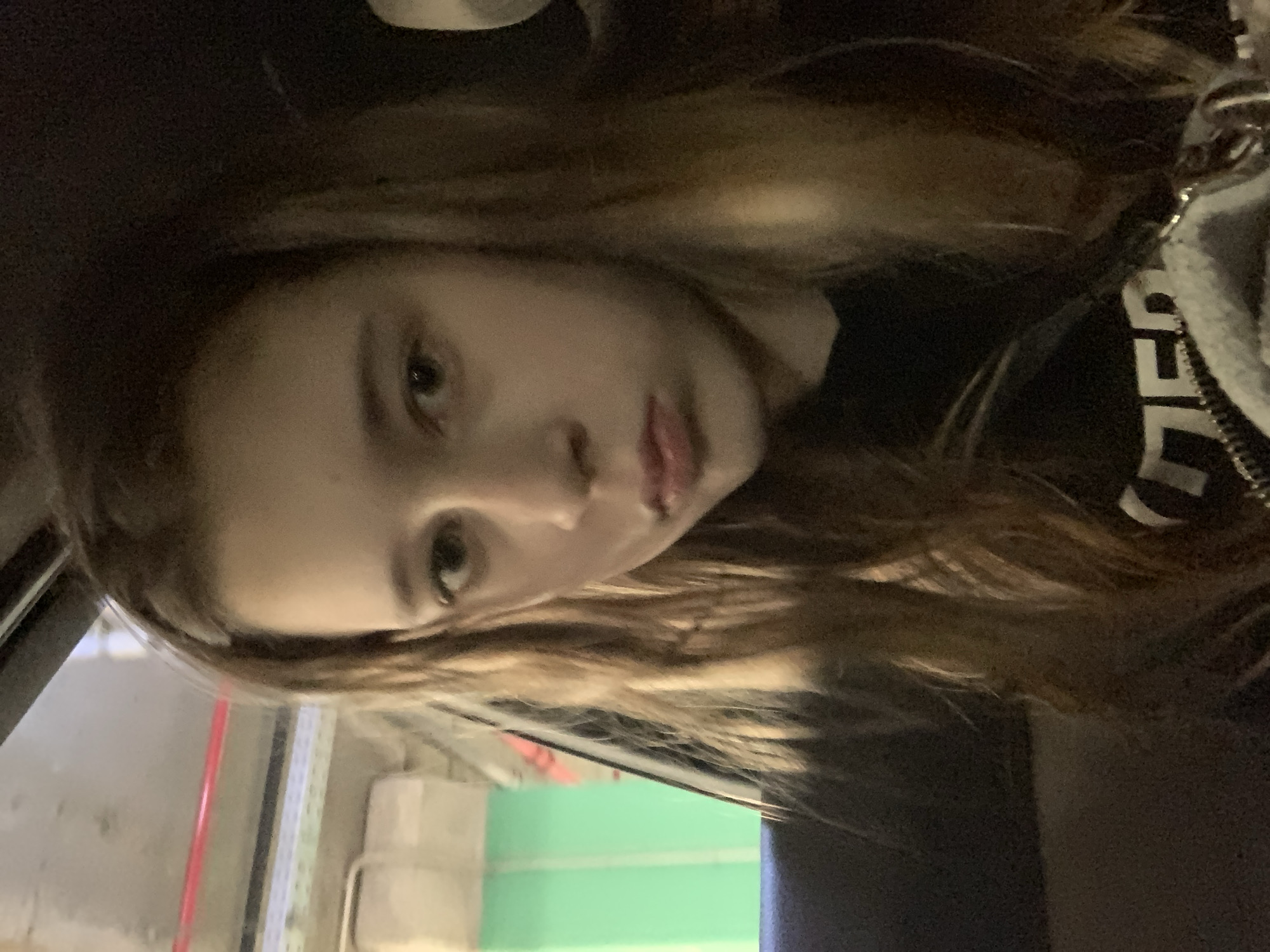 A young person with light skin and light brown hair is in the foreground, facing the camera. They appear to be inside a vehicle, possibly a car, given the door frame visible on the left. Outside the window, a blurred view of what appears to be an interior space with a green wall and red accents can be seen. Part of a black garment is on the subject, partly visible, with white lettering. The location in Belgrade, Serbia, cannot be definitively determined from the image.