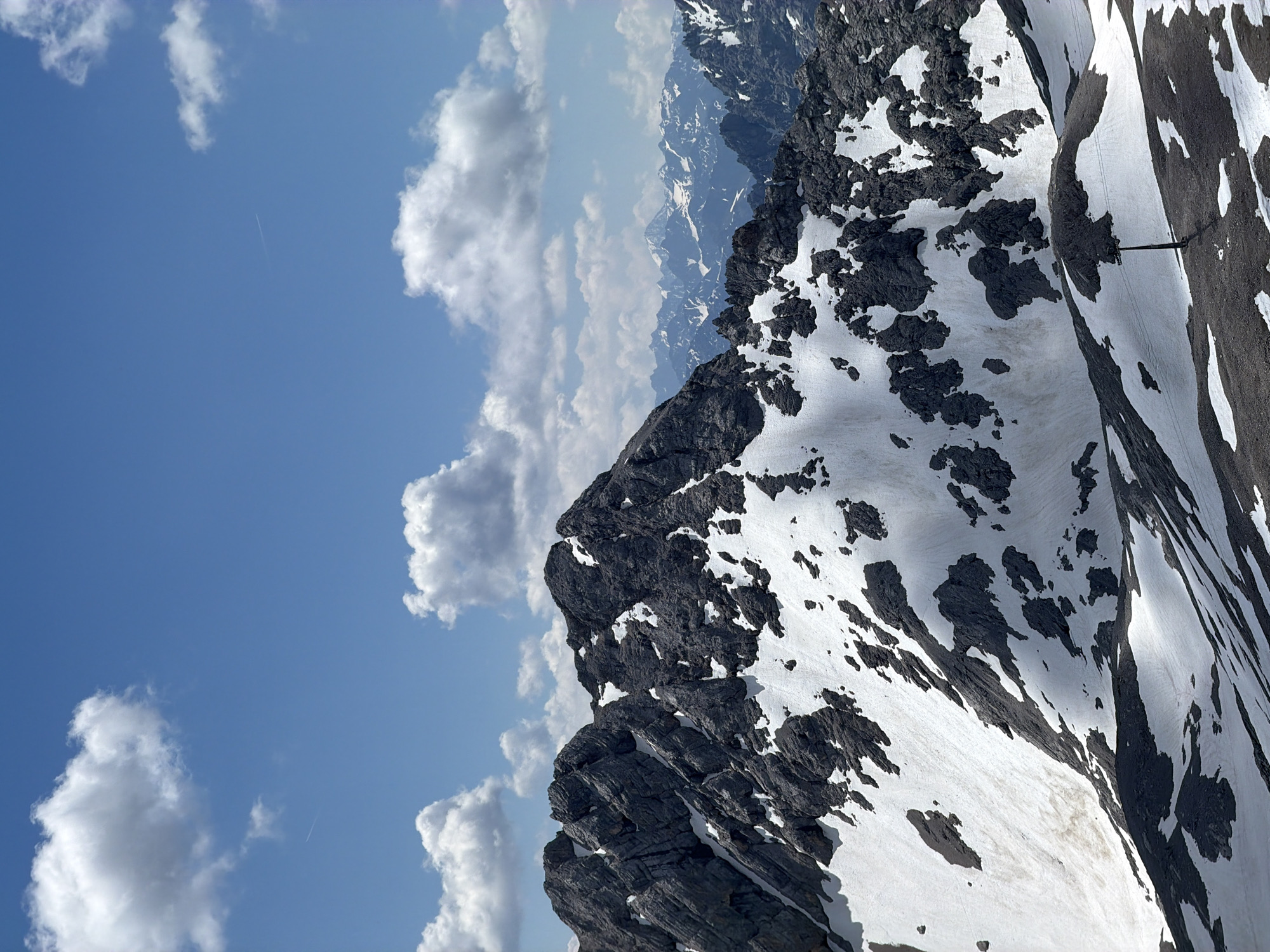 The image depicts a majestic mountain landscape under a bright, blue sky dotted with fluffy white clouds. The foreground is dominated by rugged, dark grey mountain peaks partially covered in patches of white snow. The snow is unevenly distributed, creating a stark contrast with the rocky terrain and suggesting a high altitude or recent snowfall.
In the middle ground, several other mountain ranges stretch into the distance, their peaks also dusted with snow and appearing progressively hazier due to atmospheric perspective. A lone dark pole with a wire extending from it, possibly part of a ski lift or weather station, is visible in the lower right portion of the frame, standing out against the snowy slope. The overall environment is one of natural, untamed beauty, characteristic of alpine regions.
While the image showcases a mountain scene, it does not contain any specific landmarks or elements that definitively place it within the city of Garmisch-Partenkirchen, Germany. However, the style of the mountain range and the presence of snow are consistent with the mountainous terrain surrounding Garmisch-Partenkirchen, a well-known Bavarian resort town. There are no people or animals visible in the image, and no apparent actions or interactions are taking place. The scene is a static portrayal of the natural environment.