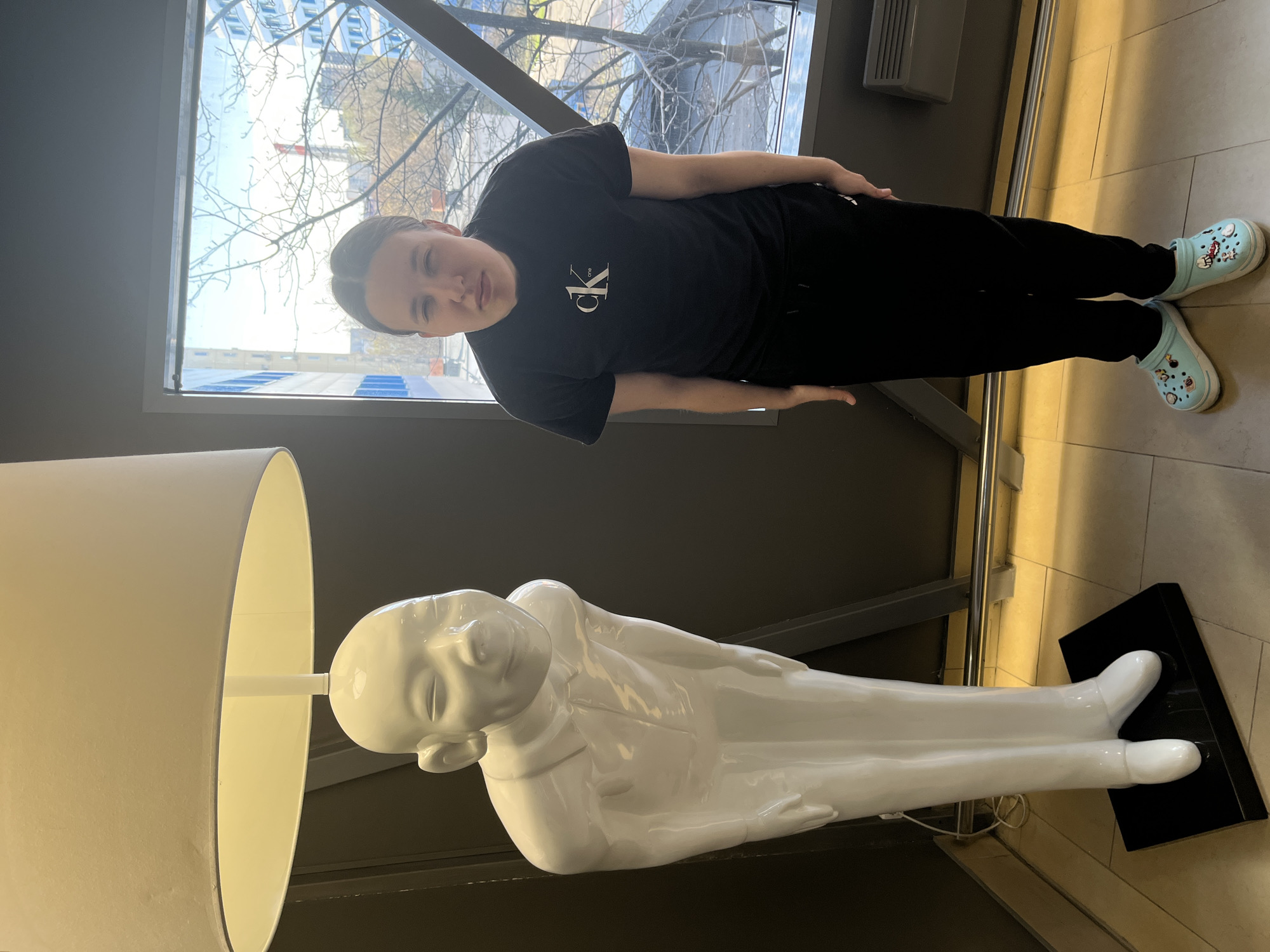 A young person stands near a large, white, recumbent figure lamp. The individual wears a black t-shirt with a "CK" logo, black sweatpants, and blue Crocs. The lamp's base is black, and a beige lampshade covers the bulb. A window behind the person shows an urban scene with trees and buildings, indicating the location is Moscow, Russia.