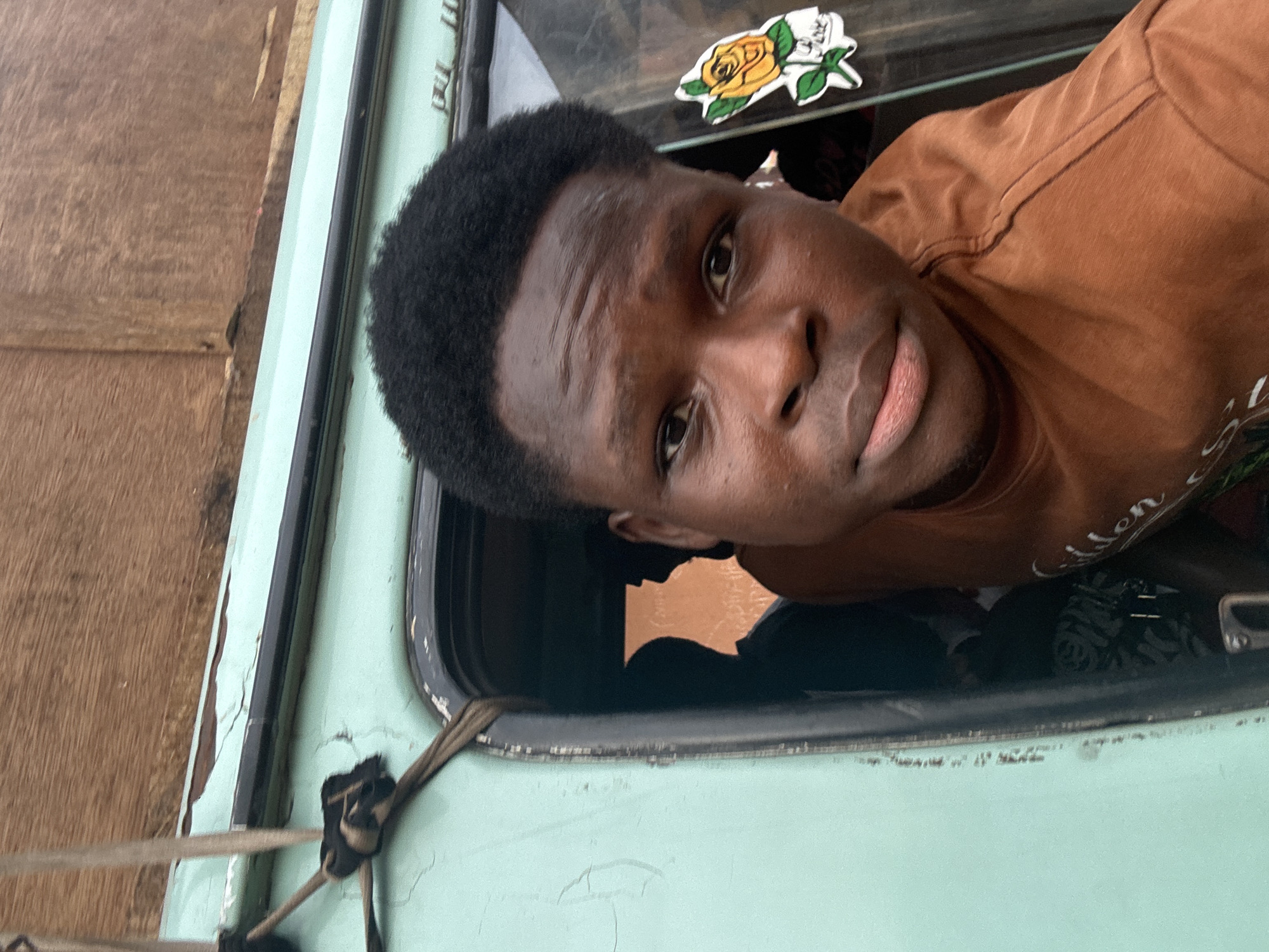 A young Black man looks out from a light green vehicle window. The paint around the window is cracked. A sticker of a yellow rose is adhered to the window. The man wears a brown t-shirt. Outside the vehicle is a wooden structure. The location is Onitsha, Nigeria.