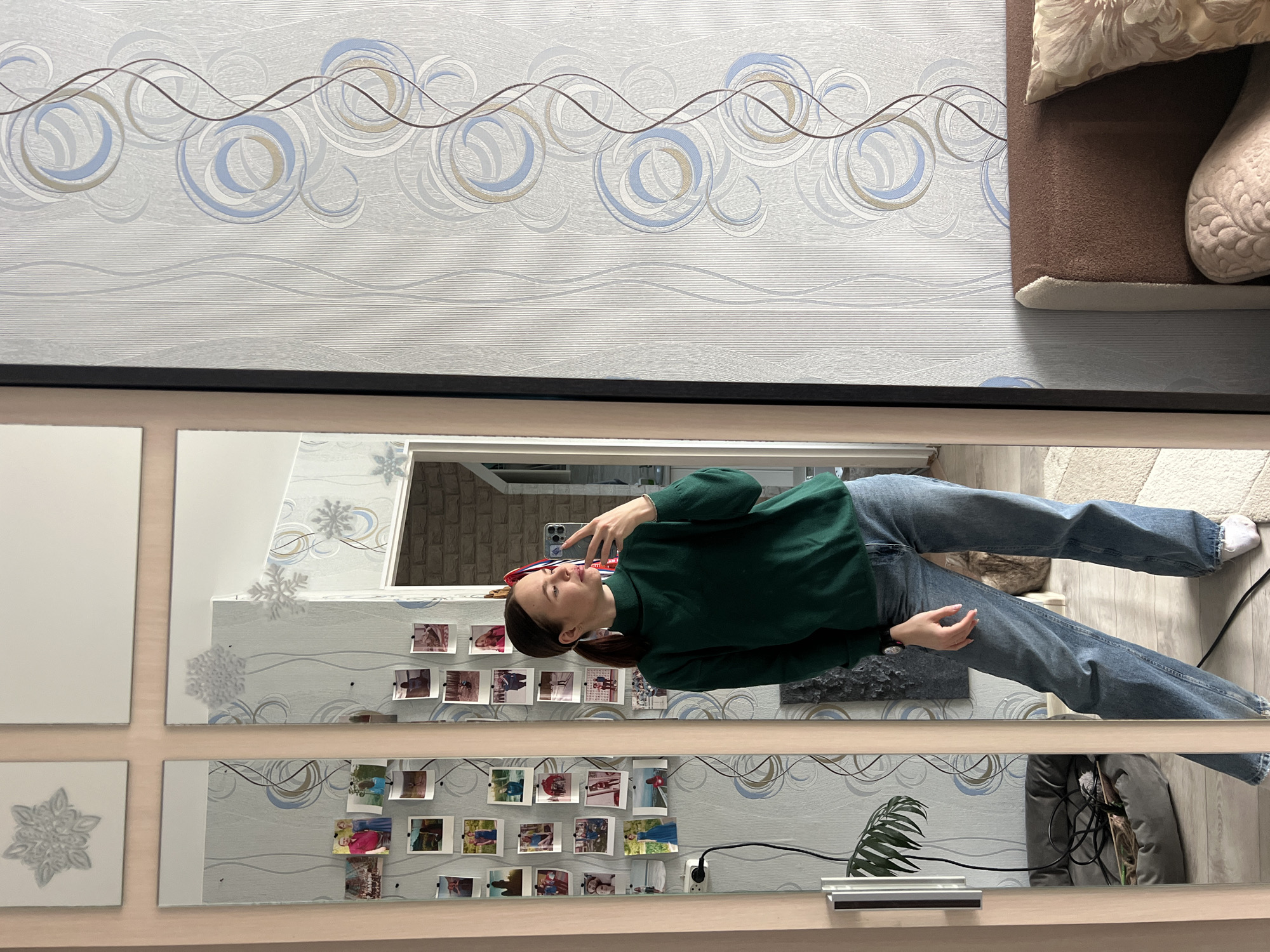 A person is taking a selfie in front of a mirror. The person is wearing a dark green turtleneck sweater and blue jeans. Behind the person is a wall with a light gray patterned wallpaper. There are small square photos taped to the wall. The mirror is segmented into multiple square panels. A couch with pillows sits in the upper right corner of the frame. The person's location may be in an apartment in Chaikovsky, Russia, judging by decor and surroundings.