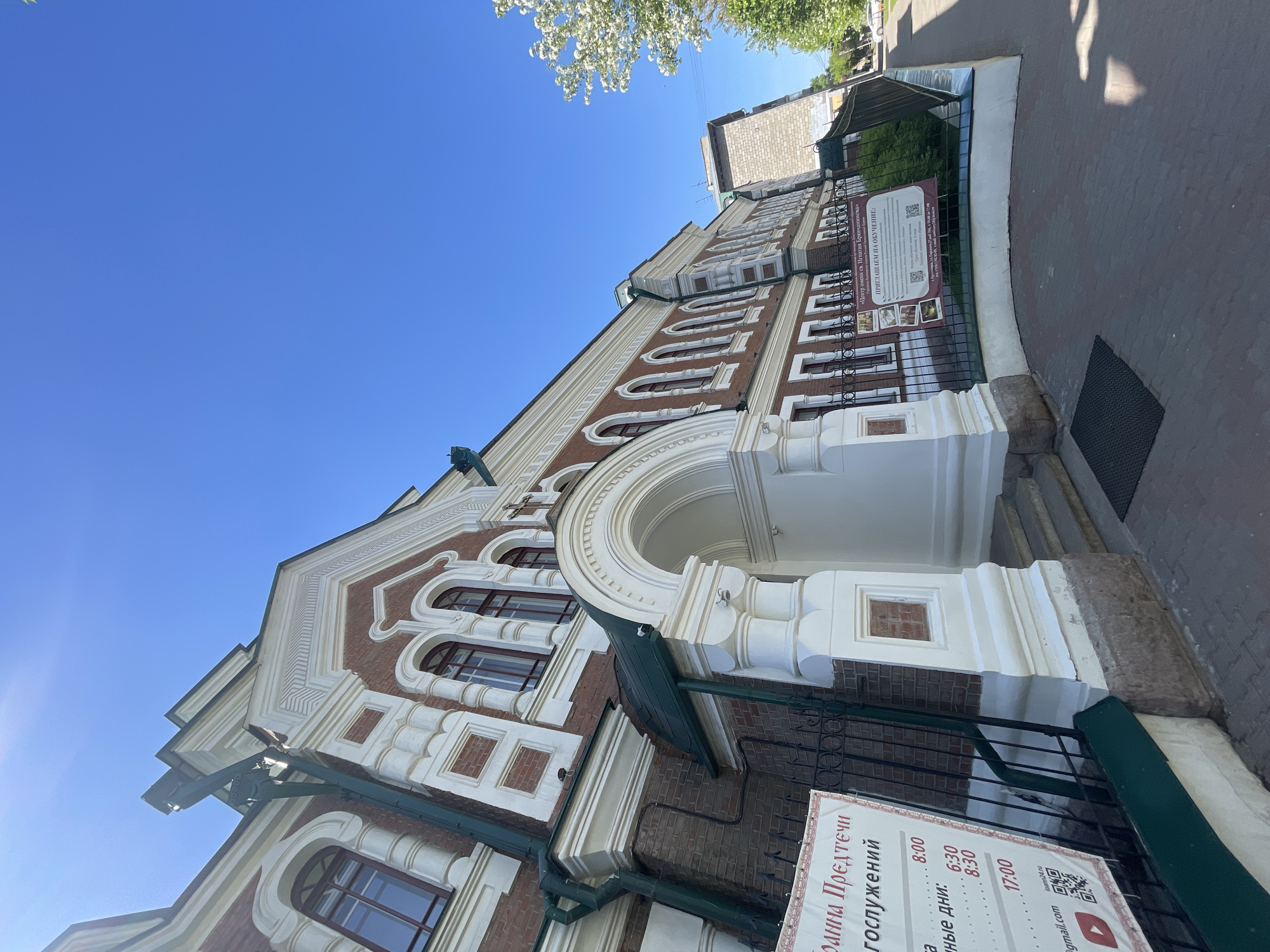 This image captures a low-angle perspective of an ornate building, likely a church or religious center, set against a clear and vibrant blue sky. The building features a red brick facade with extensive white decorative architectural elements, including arched window frames, intricate cornices, and columns surrounding a prominent arched entranceway. Green drainpipes and roof details are also visible along the structure.
In the foreground, a black metal fence runs parallel to the building, separating it from a paved sidewalk or pathway. Two signs written in Russian are attached to this fence. The sign closer to the viewer displays "Иоанна Предтечи" (John the Forerunner) and lists "богослужений" (divine services) with specific times: "8:00", "6:30", "8:30", and "17:00", indicating regular worship services.
The second sign identifies the establishment as "«Центр имени св. Игнатия Брянчанинова»" (Saint Ignatius Brianchaninov Center) and extends an invitation: "ПРИГЛАШАЕМ НА ОБУЧЕНИЕ:" (WE INVITE TO STUDY:), suggesting it functions as a religious education or cultural institution. Contact information such as "Joann24.ru", a Gmail address, a QR code, and a YouTube logo are also present on this sign.
The scene is brightly lit by natural sunlight, indicating a clear, sunny day, likely in spring or early summer, as evidenced by white blossoms and green leaves from a tree visible against the sky in the upper frame. The architectural style appears traditional, consistent with Russian ecclesiastical architecture. The building is located in Krasnoyarsk, Russia. No people or overt activities are depicted, focusing instead on the building's structure and the informational signs.