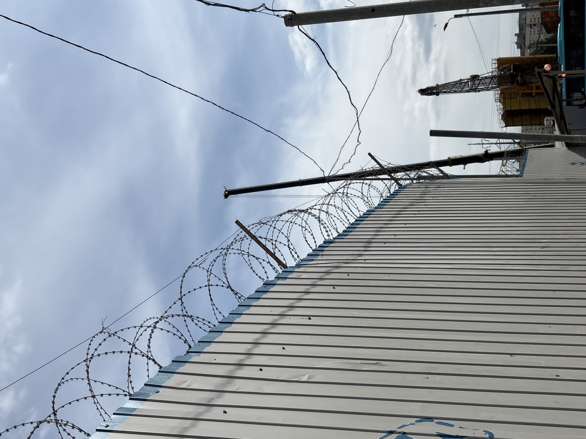 This image captures an outdoor scene, likely in an urban or industrial area within Saint Petersburg, Russia. A prominent grey corrugated metal fence or wall stretches diagonally across the frame from the bottom-left foreground towards the upper-right background. This fence is topped with multiple coils of razor wire, which are supported by several angled posts extending upwards from the barrier. A faint, blurry blue mark is visible on the lower part of the fence.
Above and beyond the fence, a cloudy sky dominates the upper portion of the image, appearing overcast or partly cloudy. Several dark utility wires crisscross the sky, and parts of utility poles are also visible. In the far distance on the right, a large yellow and black construction crane is partially visible, indicating a construction site or industrial zone. Beyond the crane, faint outlines of distant urban buildings can be discerned against the sky.
No specific activity or event is taking place; the scene appears static. The time of day is daytime, under an overcast or partly cloudy sky. The razor wire and robust fence convey a sense of security, restriction, or an industrial perimeter. No discernible text is visible in the image.