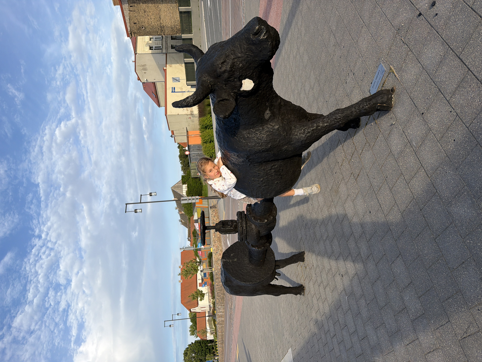 The image captures a public art installation featuring a black, abstract sculpture of a bull connected by a pipe to another bull-like form, in the city of Ventspils, Latvia. A young girl is seen lying on the back of the larger bull sculpture, resting her head on it. The sculpture is situated on a paved walkway, with buildings and streetlights visible in the background under a cloudy blue sky. The sculpture is located on Ostas iela 22, near the Ventspils Information Center.