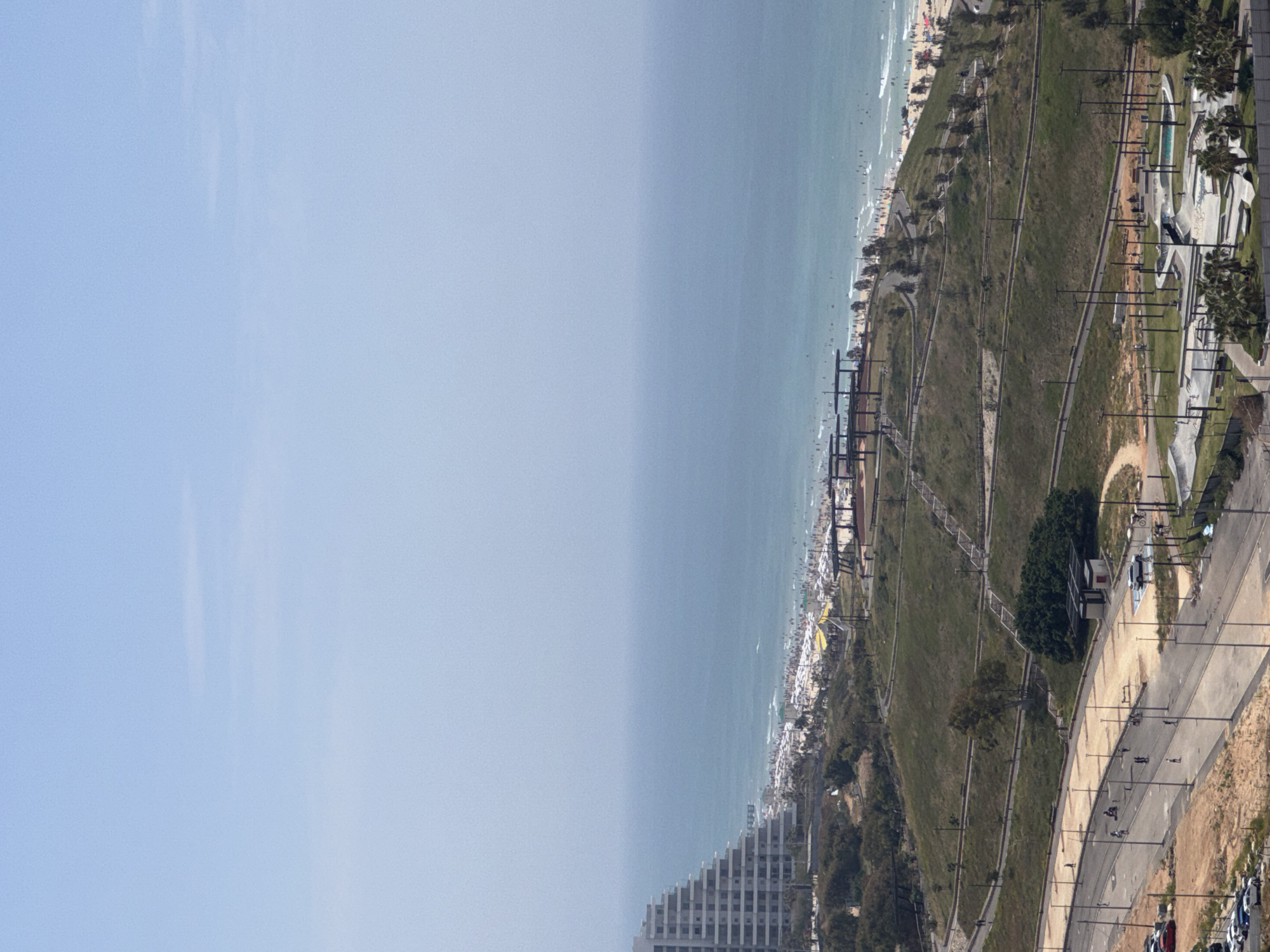 The image captures a panoramic view in Bat Yam, Israel, featuring a beachfront area. The frame is divided by the sky on the left, a large expanse of the sea in the middle, and the city's coastline on the right. A populated beach is directly next to the water. Moving inland from the beach is a green belt, with tiered walking paths. There is a road with cars and pedestrians. The area includes trees, grass, and paved walkways. The bottom portion of the photo also features a high-rise building.