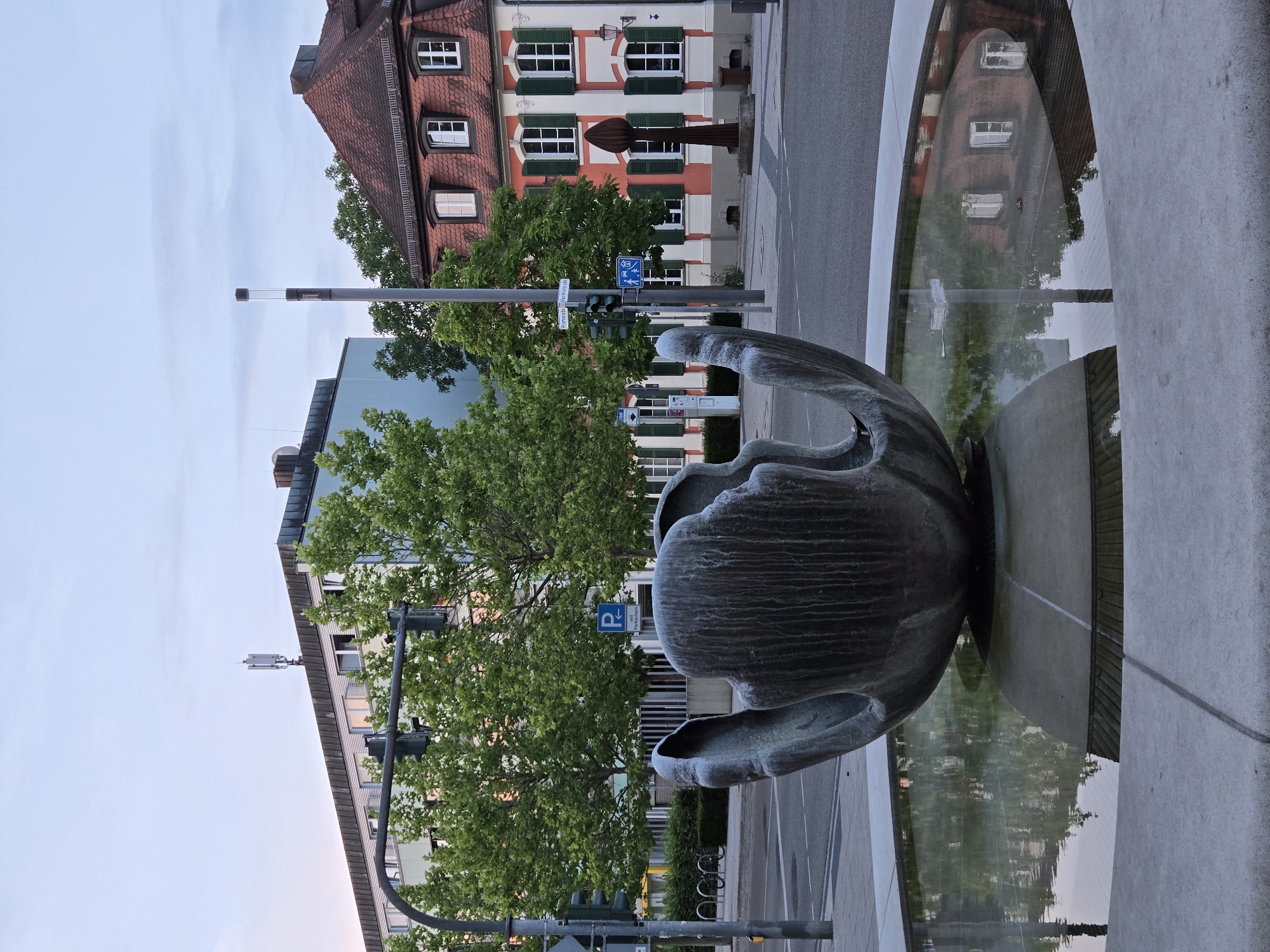 This is a ground-level, medium-shot, eye-level photograph of a modern sculpture in a fountain, situated in the city of Ansbach, Germany. The sculpture, made of dark, textured metal, resembles a large, open seed pod or a stylized flower. It is partially submerged in the still water of a circular fountain, with its upper portions rising above the water's surface. The water reflects the surrounding environment, including the sky, trees, and buildings.
In the background, on the left side of the image, is a multi-story modern building with a flat roof. A cell tower is mounted on top of this building. In front of this building, trees with lush green foliage are visible. Traffic lights and street signs are also present.
To the right of the modern building, a traditional, older building with a red tiled roof and white and red painted facade is visible. This building has multiple windows with green shutters. In front of this building, a tall, slender, dark-colored statue or monument stands.
The scene is set at dusk, with a soft, diffused light illuminating the sky, which transitions from pale blue at the top to a lighter hue near the horizon. The overall impression is one of an urban public space, likely a plaza or square, in Ansbach.