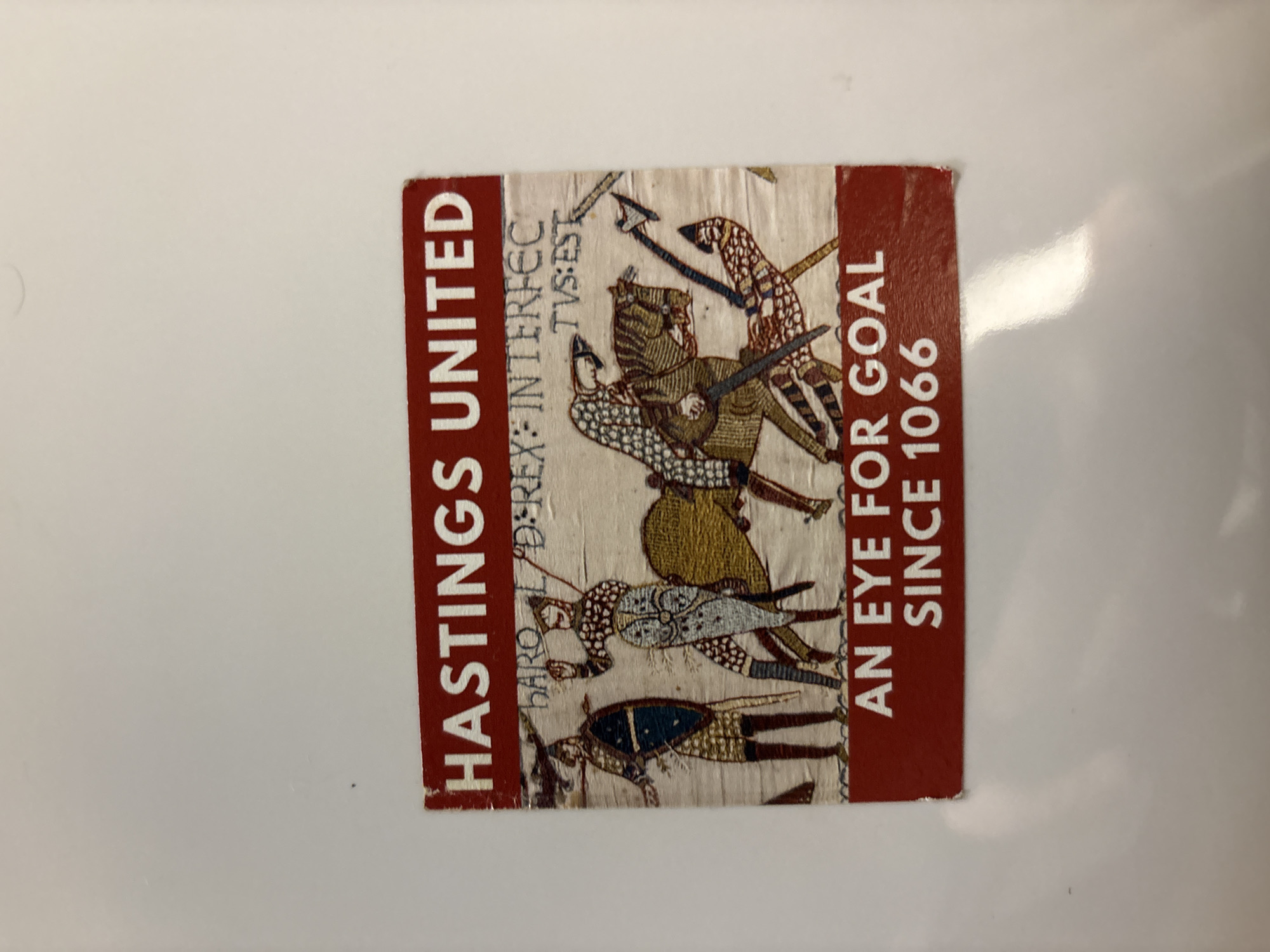 The image is a sticker for Hastings United, a football (soccer) team. The sticker is rectangular and features text on a red background alongside an image based on the Bayeux Tapestry, depicting the Battle of Hastings. On the left side, in red, is "HASTINGS UNITED." The right side, also in red, reads "AN EYE FOR GOAL SINCE 1066." Between the two red stripes is a portion of the Bayeux Tapestry, showing armed figures in battle, with a Latin inscription above them: "HARO L/D REX: INTERFEC TVS: EST." The sticker is adhered to a glossy white surface. There is no information provided to indicate it is within the city of London or anywhere in the United Kingdom specifically.