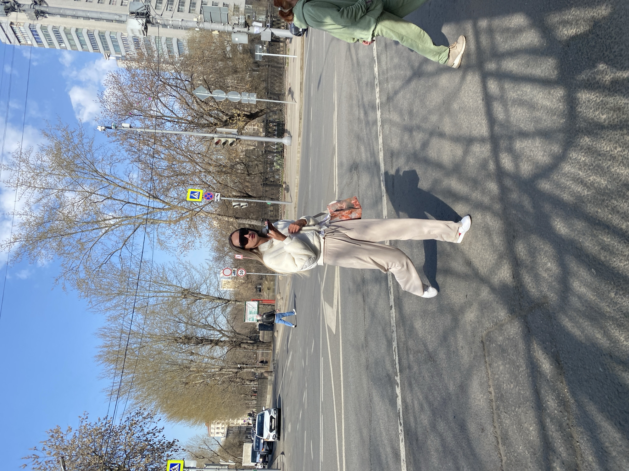 A medium shot, oriented vertically, shows two people crossing a wide, multi-lane street in Khimki, Russia. A woman in beige sweats and a white jacket stands on the crosswalk, looking at her cell phone, a purse slung on her arm. Another person, wearing a green track suit, crosses behind her and to the right. Cars are on the road to the left of the woman. Traffic lights and signs stand at intervals along the left edge of the street. Bare trees are on the left. A tall, gray building is in the upper left, above the trees. The sky is blue, with a few white clouds.