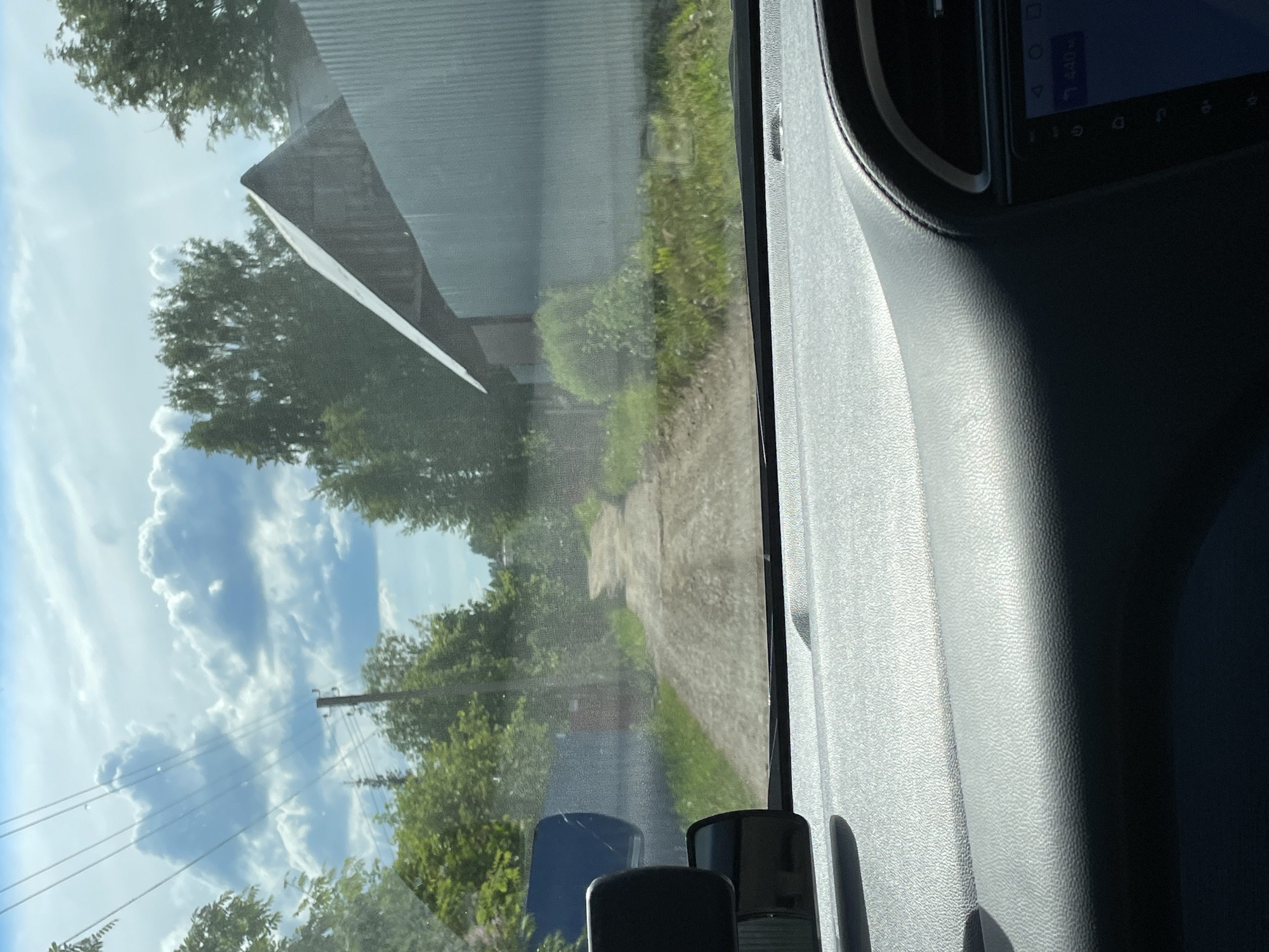 The image captures a view from inside a vehicle, looking out through the front passenger window on a bright, sunny day. The camera is oriented vertically, showing the scene as if it were viewed with the head tilted.
Outside the window, a bright blue sky with fluffy white cumulus clouds dominates the upper half of the frame. Green trees with dense foliage are scattered across the landscape, with some appearing closer and others further in the background. A utility pole with power lines is visible against the sky and trees on the left side. Running parallel to the vehicle is a dirt or gravel path, flanked by green grass and more trees. A building with a dark, corrugated metal roof is partially visible in the distance, appearing behind the trees.
Inside the vehicle, the textured gray plastic of the car's dashboard and door panel is visible on the right side, mostly in shadow, indicating strong sunlight from the left. An infotainment screen or a similar digital display is partially visible in the upper right corner, showing some interface elements and the number "440". Several black objects, possibly accessories or parts of the car's interior, are seen in the lower right portion of the image. Reflections of the car's interior are faintly visible on the window glass, blending with the exterior view.
The scene depicts a daytime setting, likely mid-day given the brightness and cloud formation, in a rural or suburban area identified as Пятый километр, Russia.