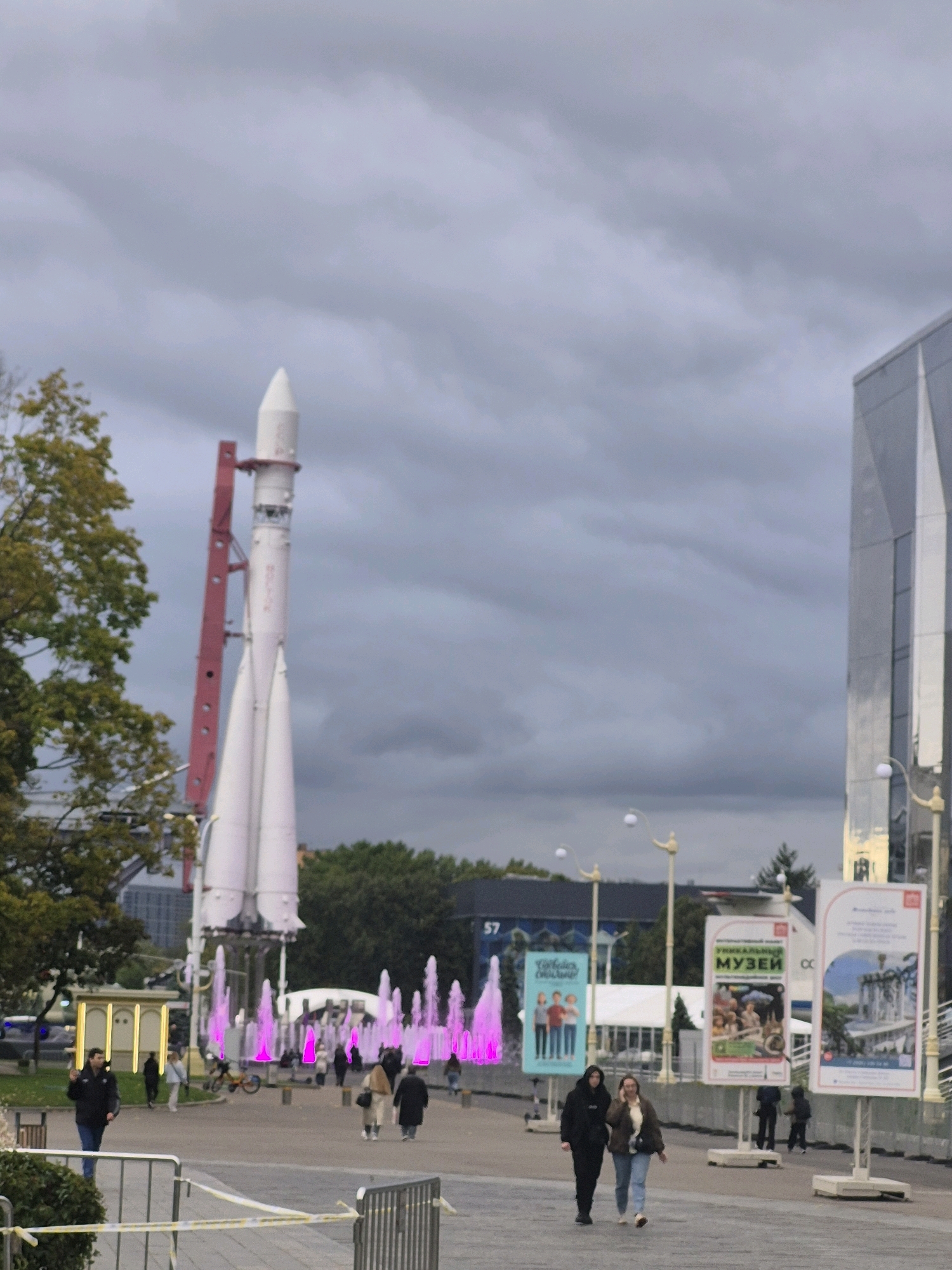 The image depicts an outdoor scene in Moscow, Russia, likely at an exhibition or park. In the foreground, a paved walkway is visible, with a man walking towards the camera from the left. Further back, several people are strolling, some with shopping bags. To the left, a large white rocket stands tall, supported by a red structure. Pink illuminated fountains are present in front of the rocket. In the background, modern buildings with reflective facades are visible, alongside trees and streetlights. Several large posters are displayed on stands, featuring images and text in Russian, suggesting an event or attraction. The sky is overcast with dark clouds.