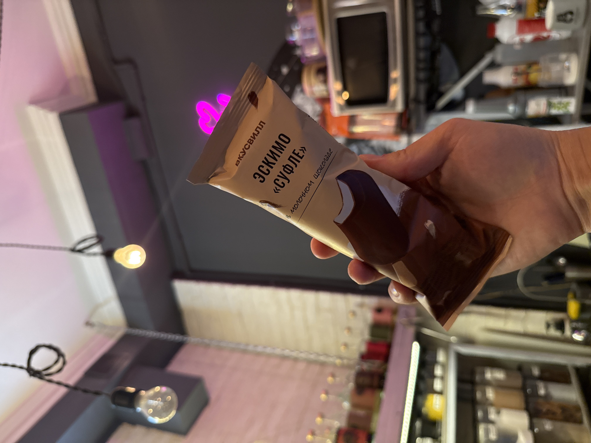 A hand holds a chocolate ice cream bar with a wrapper that reads "ЭКСКИМО «СУФЛЕ»" in Russian, which translates to "Eskimo Soufflé." The wrapper also has text that says "в молочном шоколаде," meaning "in milk chocolate." The ice cream appears to be dipped in dark chocolate, with a white base visible at the top.
The background suggests an indoor setting, possibly a bar or cafe, with shelves of bottles and jars visible. There are also decorative lights, including a string of small, illuminated bulbs and a pink neon heart shape. The lighting is dim, with a warm glow from the bulbs. The overall impression is a casual, modern environment. There are no people fully visible, only the hand holding the ice cream. The time of day cannot be determined, but the indoor lighting suggests it could be evening or night.