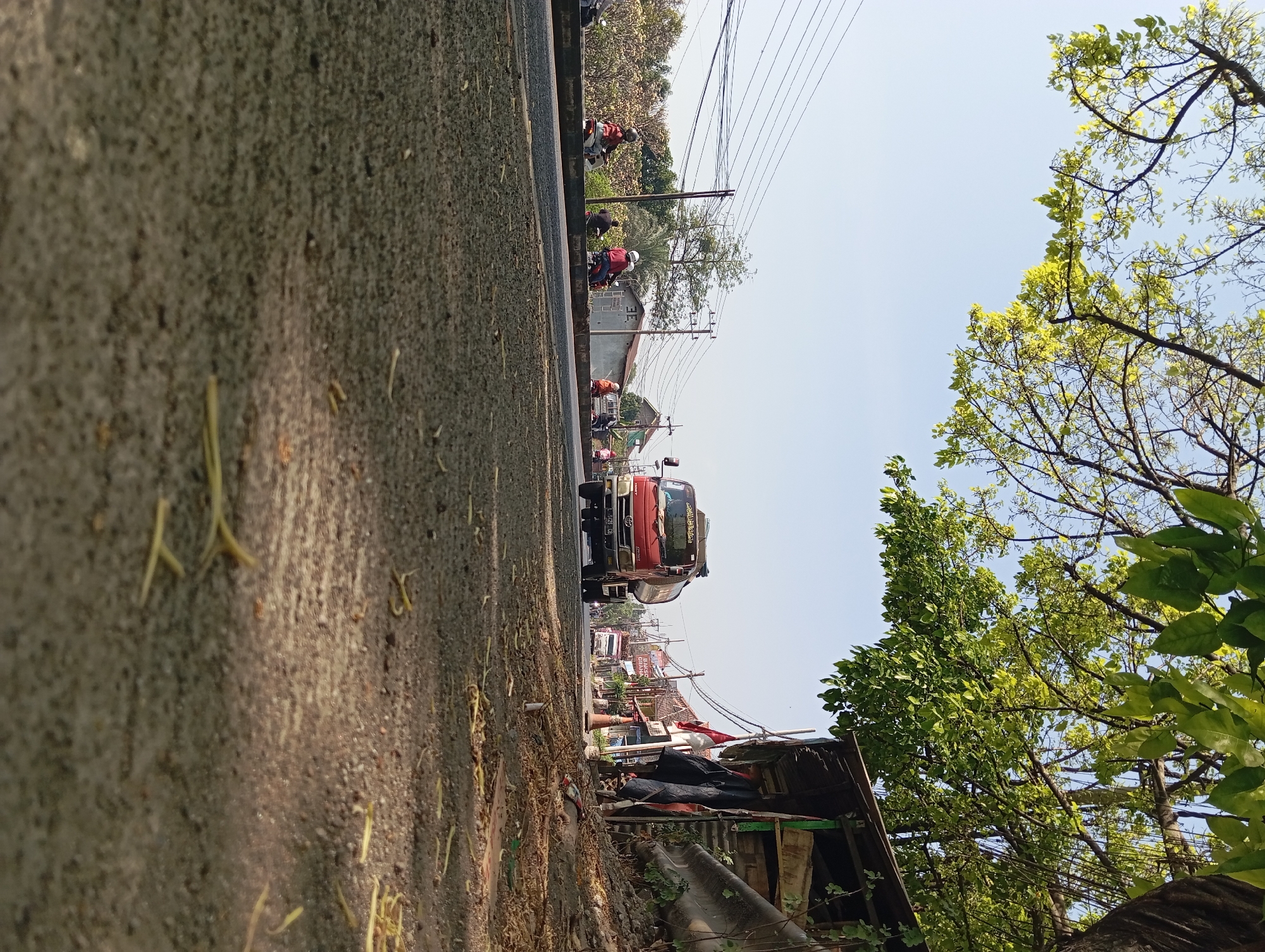 The image depicts a roadside scene in Kepulungan, Indonesia. A red tanker truck is driving on a paved road, with a clear blue sky and green trees in the background. Several motorcycles are also visible on the side of the road. A small, makeshift shelter with a corrugated metal roof stands on the right side of the image. The ground is covered with asphalt and scattered debris, including leaves and twigs. The overall scene suggests a rural or semi-urban environment with ongoing traffic.