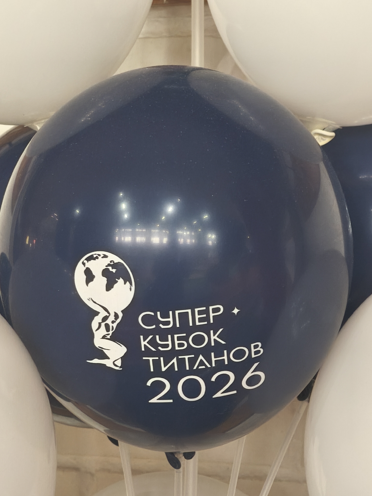 A close-up shot depicts a dark blue balloon surrounded by white balloons. The blue balloon features a white image of Atlas holding up the globe, along with the white text "СУПЕР КУБОК ТИТАНОВ 2026". The scene is indoors. Based on the text on the balloon in Russian and reflection of lights from the ceiling in the blue balloon, it is likely that it's in Saint Petersburg, Russia.