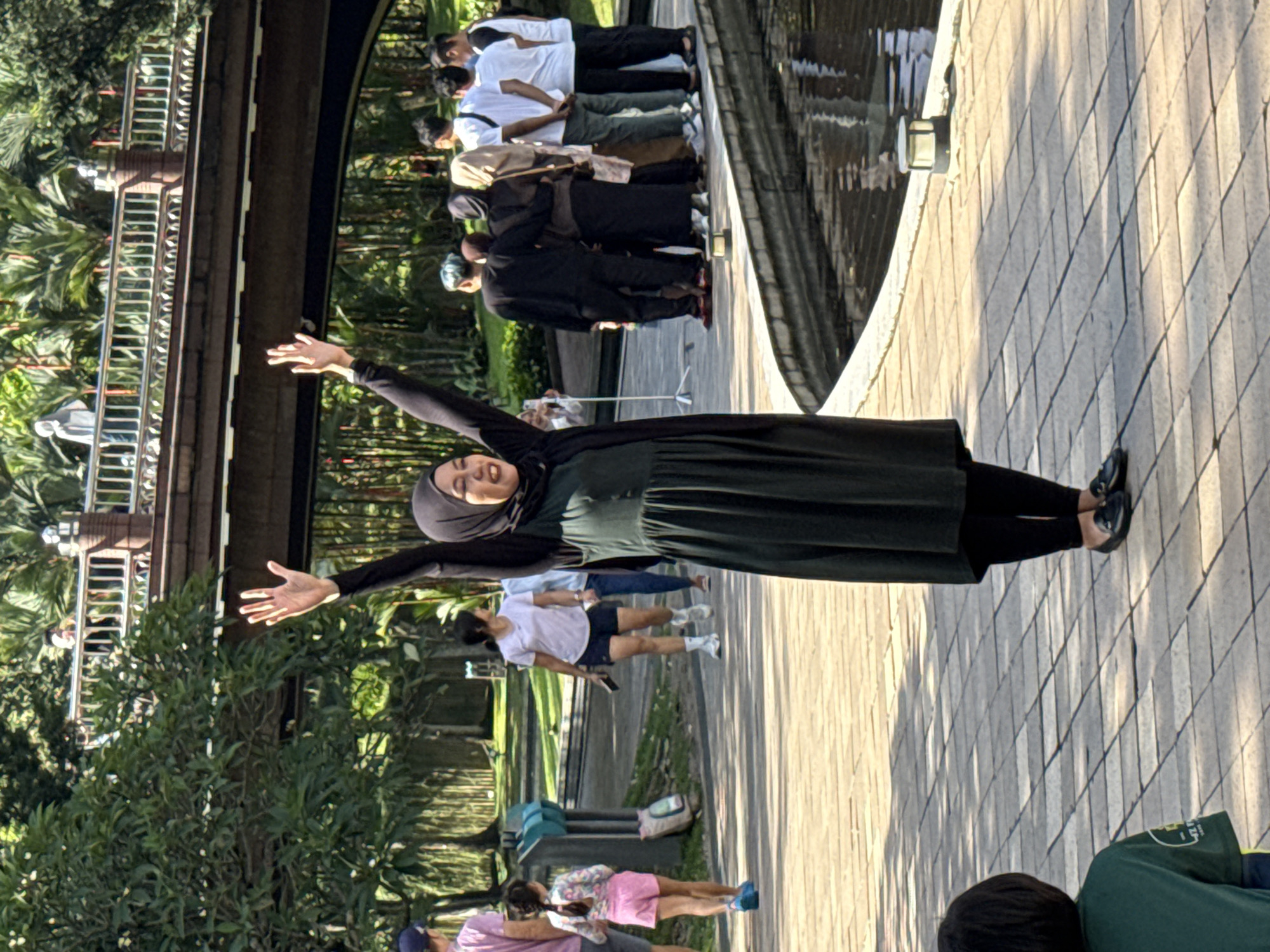 The image captures a scene in Kuala Lumpur, Malaysia. A woman wearing a hijab stands on a paved pathway with her arms outstretched. In the background, a group of people are lined up along the pathway near a structure resembling a bridge or elevated walkway. Trees and greenery surround the area. The pathway is made of light-colored paving stones. Other individuals are also visible on the pathway, some seemingly engaged in casual activities.