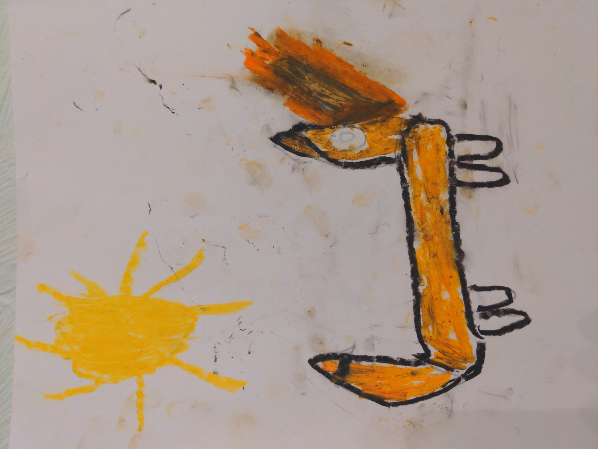 A child's drawing on white paper features an abstract representation of a person's head or face, rendered primarily in yellow and orange crayon. The face is angular and stylized, with a tuft of brown and orange "hair" atop the head. Two black shapes resembling ears or appendages are on the right side of the face. To the left of the face is a crude drawing of a yellow sun. The paper shows smudges and marks, indicating it has been handled. There is no contextual information suggesting it is located in Moscow, Russia.