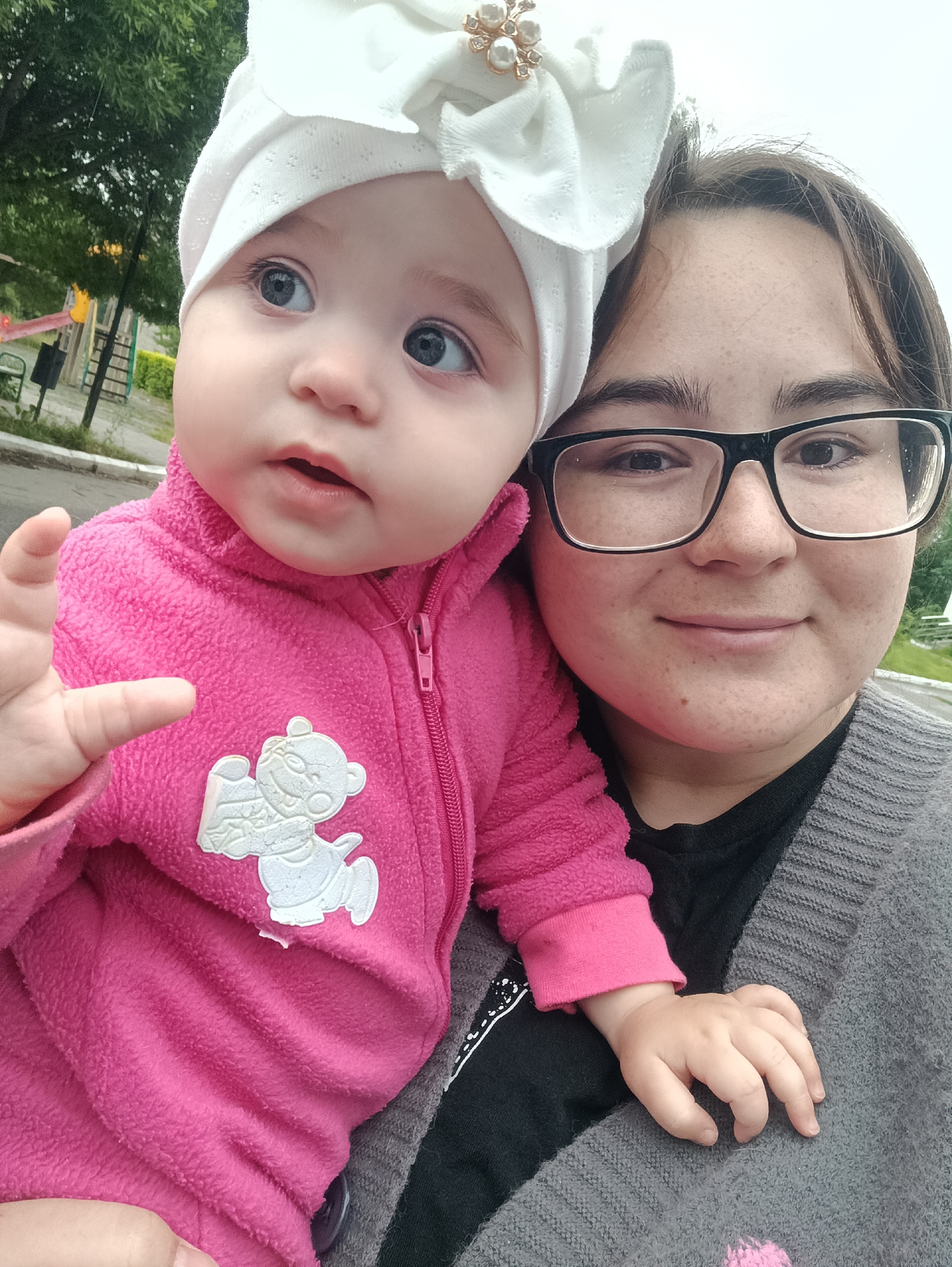 This is a selfie of a young woman holding a baby. The woman is wearing glasses and a gray cardigan over a black shirt. The baby, swaddled in a pink fleece jacket and a white bonnet adorned with pearls, looks upwards with wide blue eyes. The baby's hand is reaching out towards the camera. In the background, there's a hint of a playground with green trees and a cloudy sky, suggesting an outdoor setting. The overall mood appears affectionate and candid, likely captured during a casual outing.