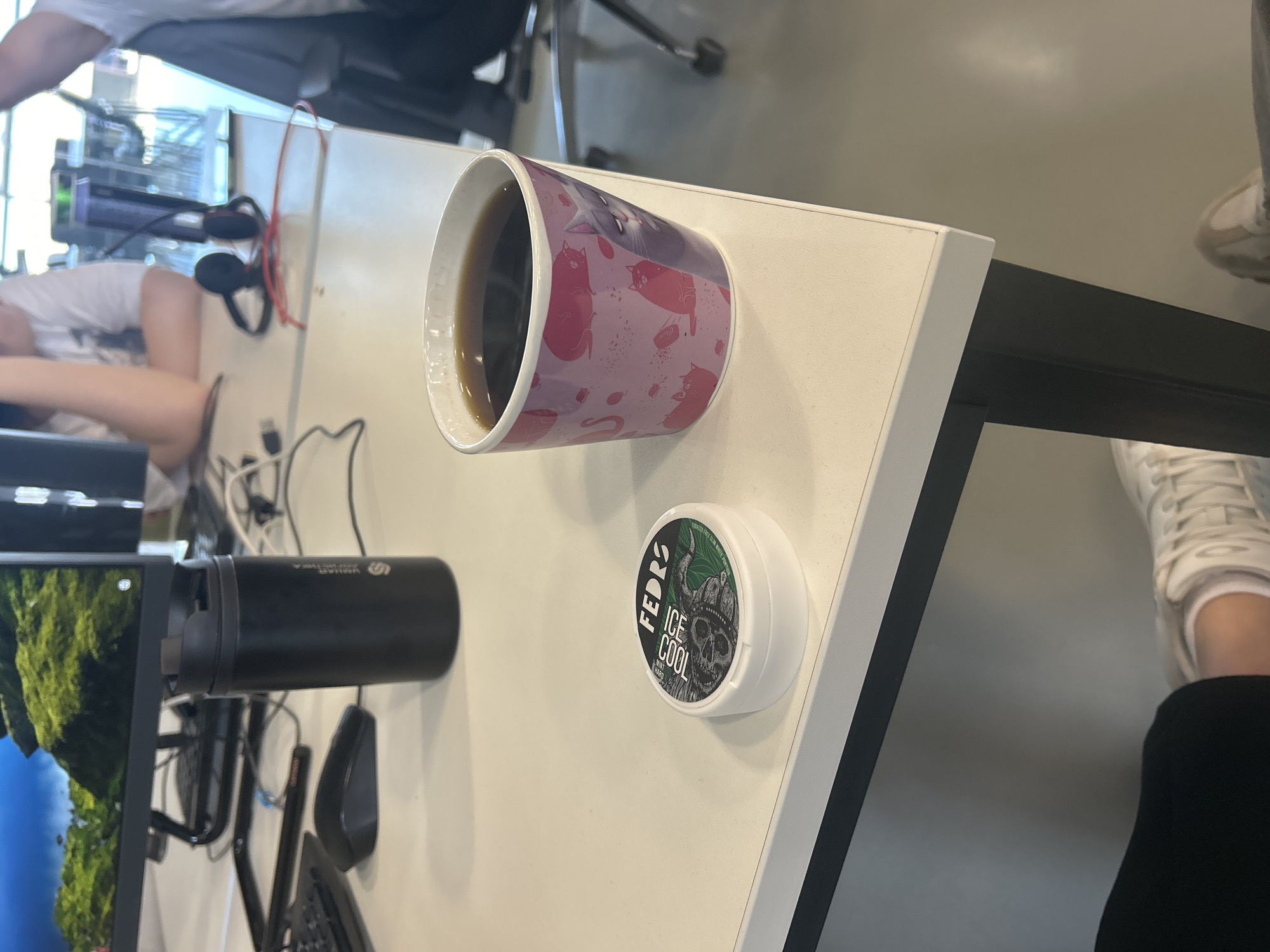 A white desk in an office environment. On the desk, there is a black computer monitor displaying a natural landscape, a black computer mouse, a black thermos, and a white mug filled with a dark liquid, adorned with pink cat illustrations. To the left of the mug, there is a small white container with a label indicating "FEDRS ICE COOL". In the background, two individuals are visible, and a pair of headphones rests on the desk. The scene appears to be an office workspace. The presence of a monitor, keyboard, and desk suggests an indoor office setting. The general aesthetic and items visible do not provide specific clues to pinpoint the exact location within the city of Perm, Russia.