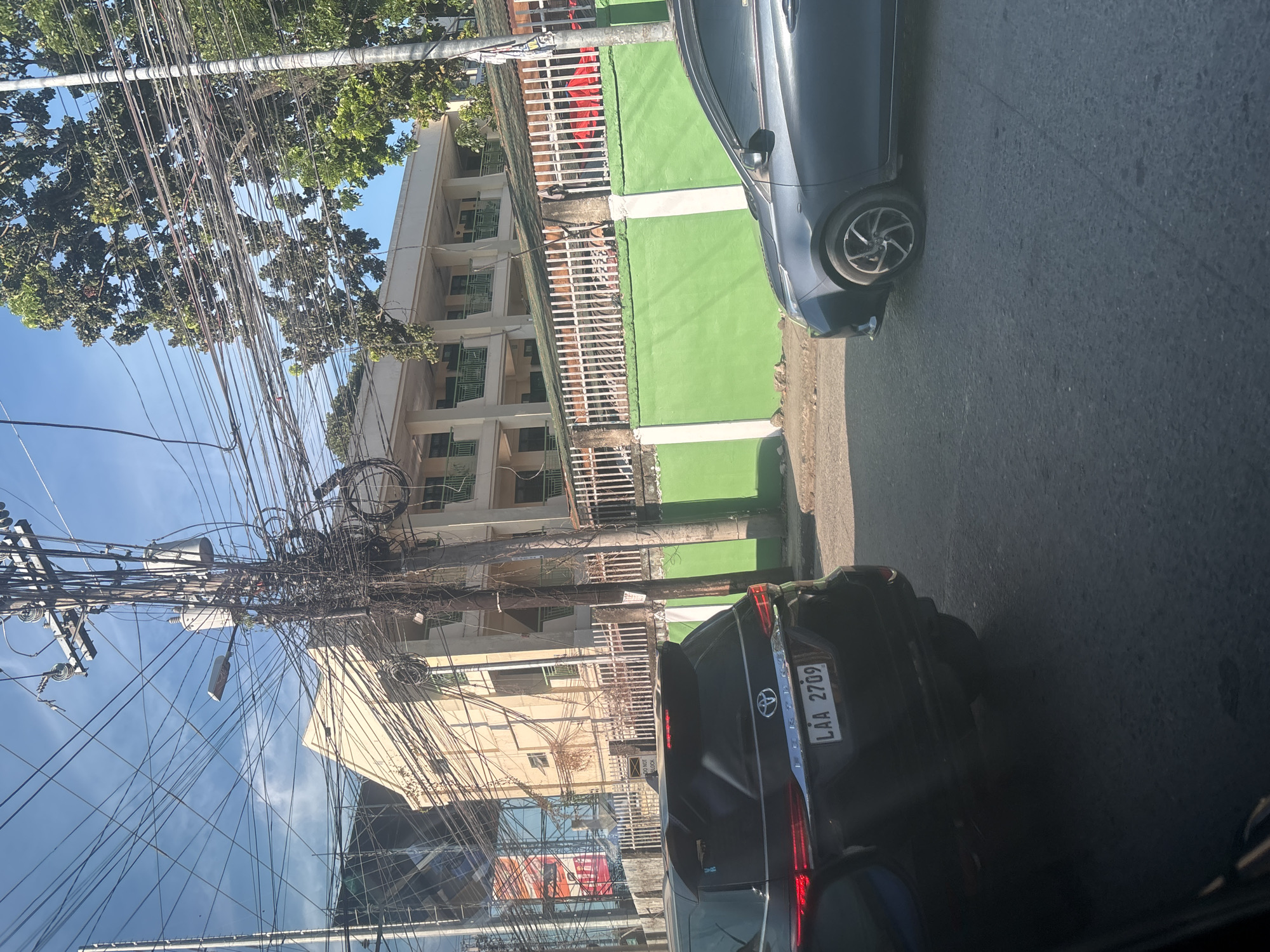 The image depicts a street scene in Davao City, Philippines. In the foreground, a black SUV with the license plate LAA 2709 is visible on the left. To the right, a portion of a dark-colored sedan is seen. Behind the vehicles, a light-colored, multi-story building with multiple balconies and green railings stands. The building is partially obscured by a dense network of electrical wires and utility poles that dominate the upper portion of the image. A bright green wall with a white fence on top runs along the front of the building. Trees with lush green foliage are also present, interspersed with the wires. The sky is clear and blue. The overall impression is of an urban environment with visible infrastructure, including street-level traffic and overhead utility lines.