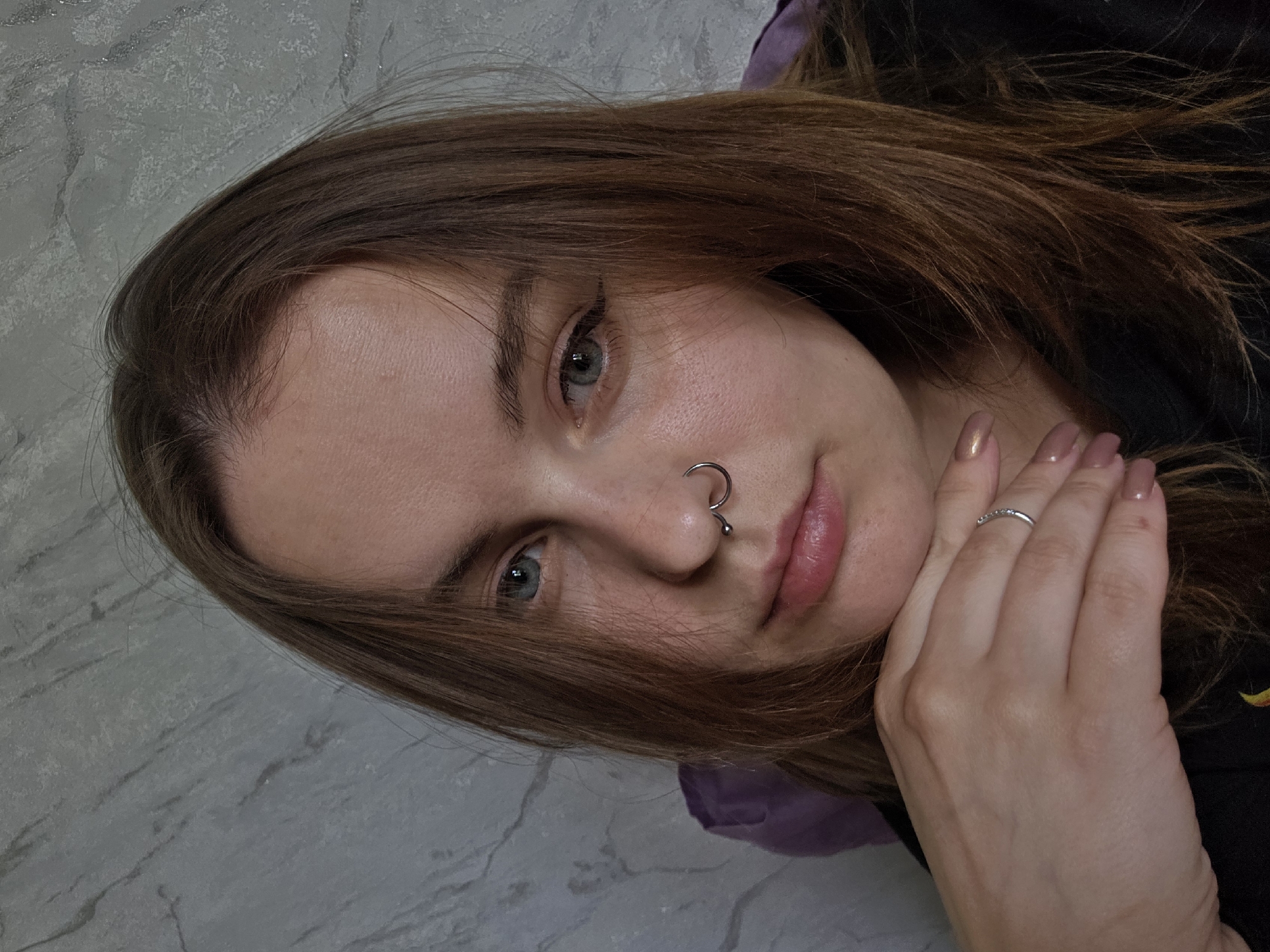 A close-up, slightly angled portrait of a young woman with light brown hair, pale skin, and light-colored eyes. She is wearing a silver ring on her finger and a silver hoop nose piercing. Her fingernails are manicured with a neutral color. Her hand is positioned near her face. The background consists of a patterned wall, a purple fabric, and a black garment. The location is unknown.