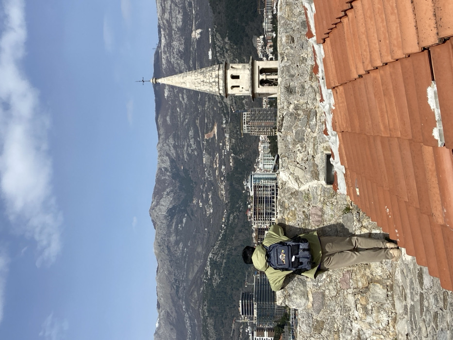 A person is observed from behind, standing on a textured stone wall, gazing across the city of Budva, Montenegro, towards a mountainous backdrop. The individual wears an olive-green hooded jacket, tan trousers, and a grey backpack with a multicolored emblem. To the immediate right, a section of a red-tiled roof is visible below the wall. The cityscape features multiple modern, multi-story buildings, some under active construction, alongside older structures. Prominently positioned to the right is a distinct light-colored stone church spire with a conical top and a cross, identifiable as the Church of Saint Ivan, featuring two levels of arched openings. The background reveals a rugged mountain range under a clear blue sky with sparse white clouds.