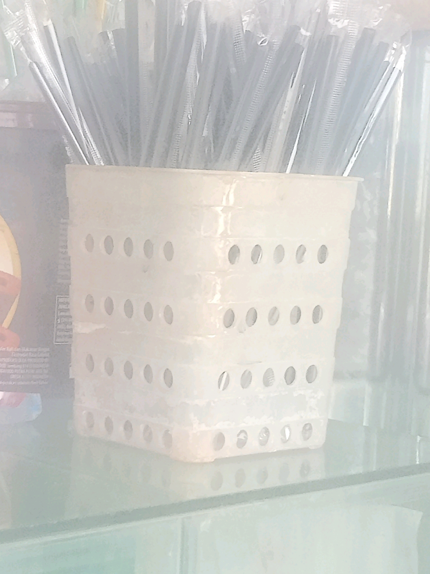 In Penusupan, Indonesia, a white, perforated plastic holder containing numerous individually wrapped black drinking straws stands on a glass shelf. The straws are bundled together, with their tops extending above the holder. The holder has multiple rows of circular perforations. To the left of the holder, a portion of packaging with text and a circular graphic is visible. The overall lighting is bright and diffused, creating a hazy effect.