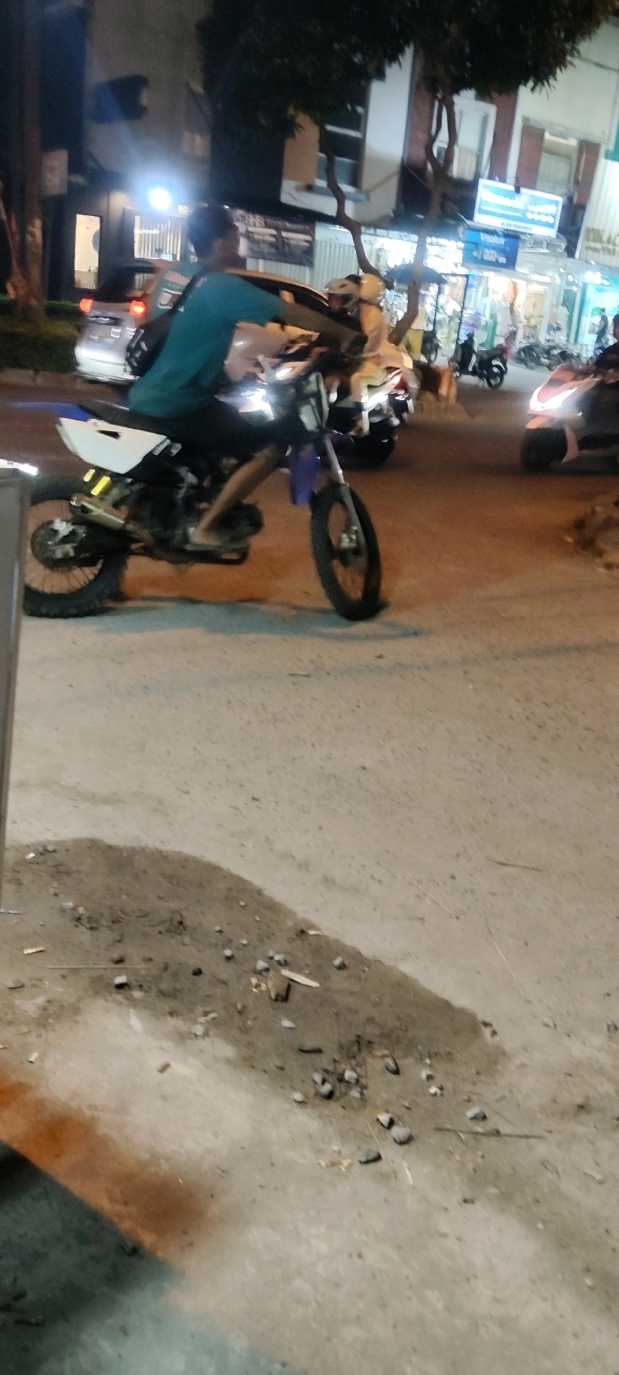 The image shows a street scene at night in Cibatu, Indonesia. A person is riding a dirt bike. The rider is wearing a teal shirt and black pants and is barefoot. The dirt bike is white with yellow reflectors on the rear fender. The bike has a silver exhaust pipe and knobby tires. In the background, there are buildings with lights on, and other motorcycles are visible. The street surface is paved and appears somewhat worn. There is a patch of dirt and debris on the right side of the road. The lighting suggests artificial illumination from streetlights and building lights.