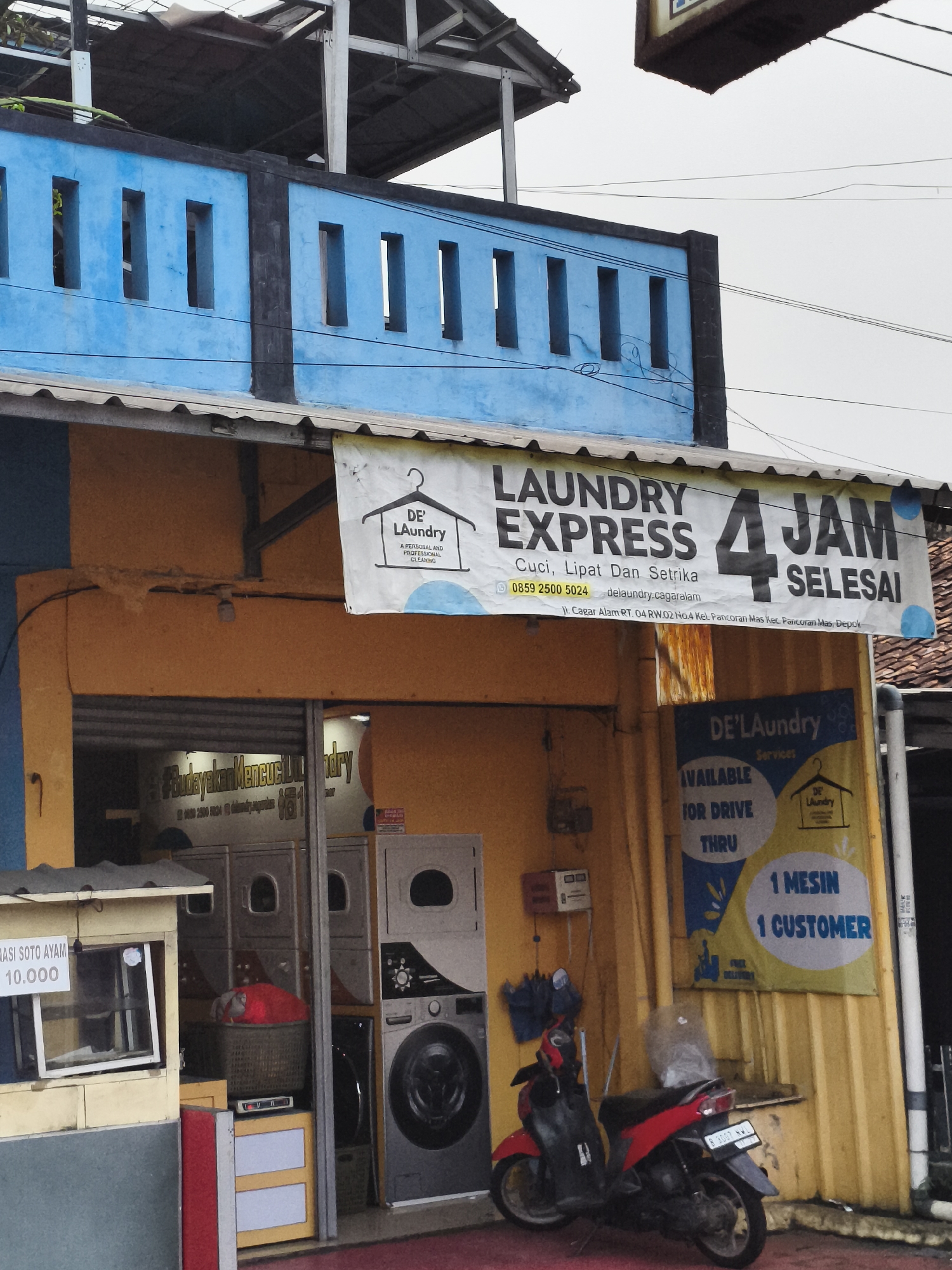Here's a detailed description of the image:
The image shows the exterior of a laundry business named "DE' Laundry Express" located in Depok, Indonesia. A banner above the entrance advertises "Laundry Express 4 Jam Selesai" (Laundry Express 4 Hours Ready) along with the services "Cuci, Lipat Dan Setrika" (Wash, Fold, and Iron). A phone number (0859 2500 5024) and the location (Jl. Cagar Alam RT. 04 RW.02 No.4 Kel. Pancoran Mas Kec. Pancoran Mas, Depok) are also listed. Another smaller sign in front indicates "Available for Drive Thru" and promotes a "1 Mesin 1 Customer" (1 Machine 1 Customer) policy.
Inside the establishment, several washing machines are visible. A red and black scooter is parked outside, next to the laundry shop. The building itself has a yellow and blue facade. To the left of the laundry, there is a small food stall selling "MASI SOTO AYAM 10.000", suggesting that the laundry shop is located in a busy or residential area where food vendors are common. The blue part of the laundry shop has rectangular hollow shapes.