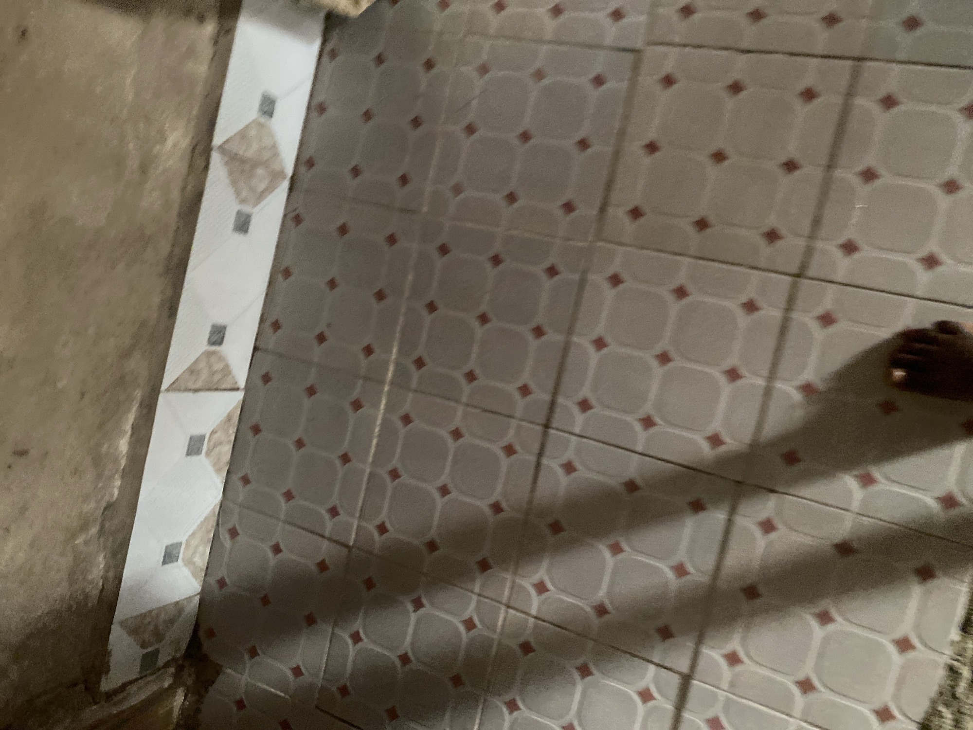 A low-angle shot captures a tiled floor in Jauro Yinu, Nigeria. The floor is covered in square tiles with a pattern of rounded shapes and small red diamond accents in the center of each tile. A strip of decorative trim tiles runs along the wall, featuring geometric patterns of white, grey, and beige. The wall above the trim appears to be rough, unfinished plaster or concrete. A person's foot and lower leg are visible in the lower right corner of the frame, casting a shadow on the floor. The lighting suggests an indoor environment, possibly a room or hallway.