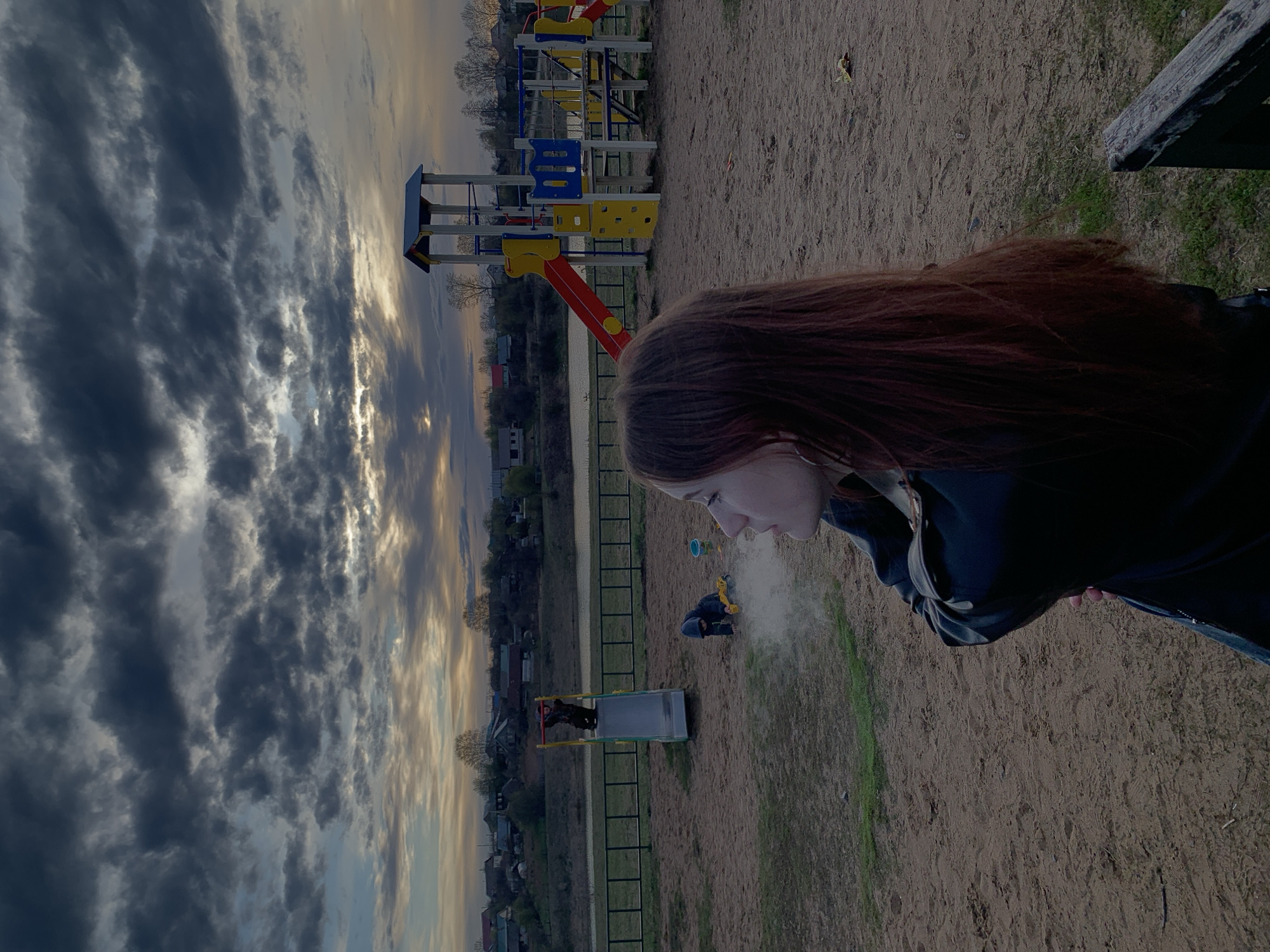 The image shows a young woman with long auburn hair standing in a sandy area in Фоки, Russia. She is wearing a dark jacket. In the background, there is a playground with slides and climbing structures. A body of water can be seen behind the playground. A person is on their knees working on the sand near the water. The sky is filled with dramatic clouds, suggesting either a sunrise or sunset.