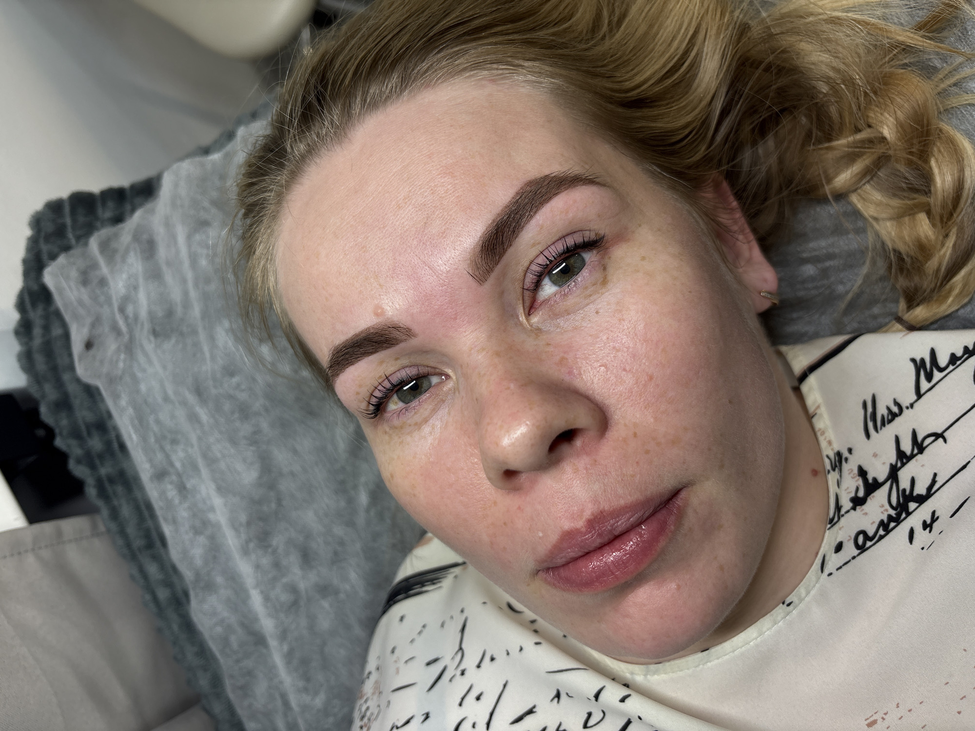 A fair-skinned woman with freckles is lying on a gray, textured surface, possibly a pillow or cushioned table, in what appears to be an indoor setting. Her blonde hair is pulled back, revealing her face. She has brown eyebrows, and her lips are lightly glossed. The text on her white top reads "Hass. M" and "Steghk." Given the text's apparent connection to Russia, the image was likely taken in the city of Perm, Russia.