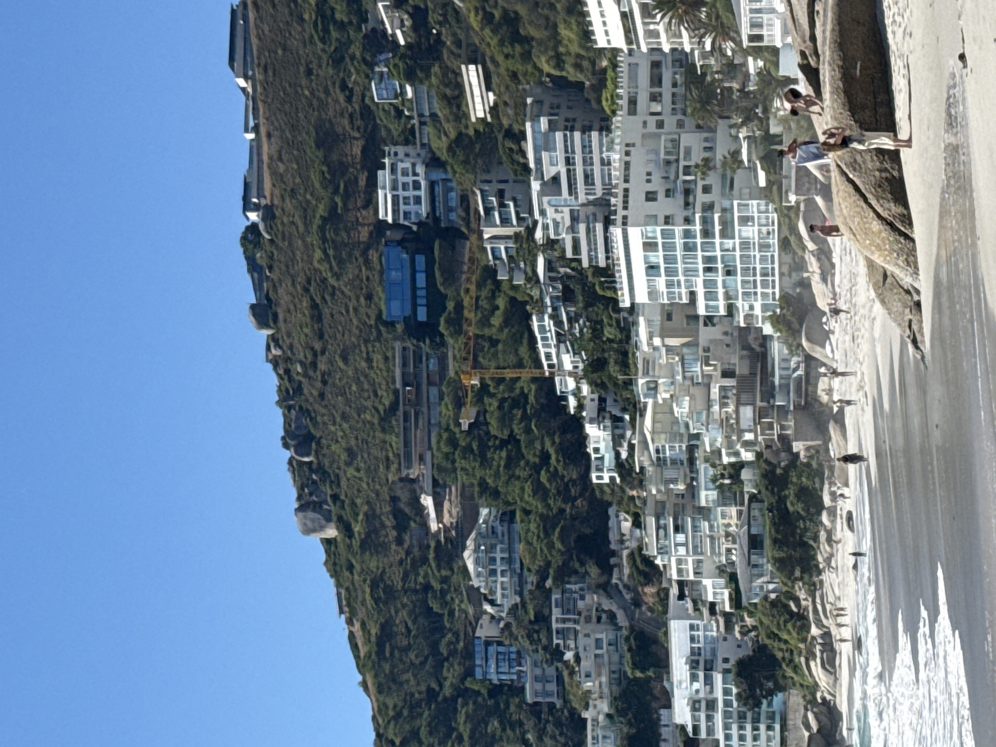 The image depicts a sunny coastal scene in Cape Town, South Africa, specifically along the Atlantic Seaboard, consistent with areas like Clifton or Camps Bay. A steep, green hillside, densely covered with vegetation and large boulders, dominates the background. Numerous multi-story, modern residential buildings with white facades and extensive glass windows are terraced up the mountainside, interspersed with trees. A yellow construction crane is visible mid-slope, indicating active development. The foreground features a sandy beach with the ocean to the left, where white waves break onto the shore. Several large, light-colored boulders are present on the beach in the immediate foreground, right of center. Approximately fifteen individuals are scattered across the beach and near the water. One person, dressed in light clothing, stands on a prominent boulder in the foreground, facing towards the ocean or the left side of the frame. Another individual is seen crouching or climbing near this boulder. Other people are walking, standing, and interacting along the sandy expanse and at the water's edge. The sky is clear blue.