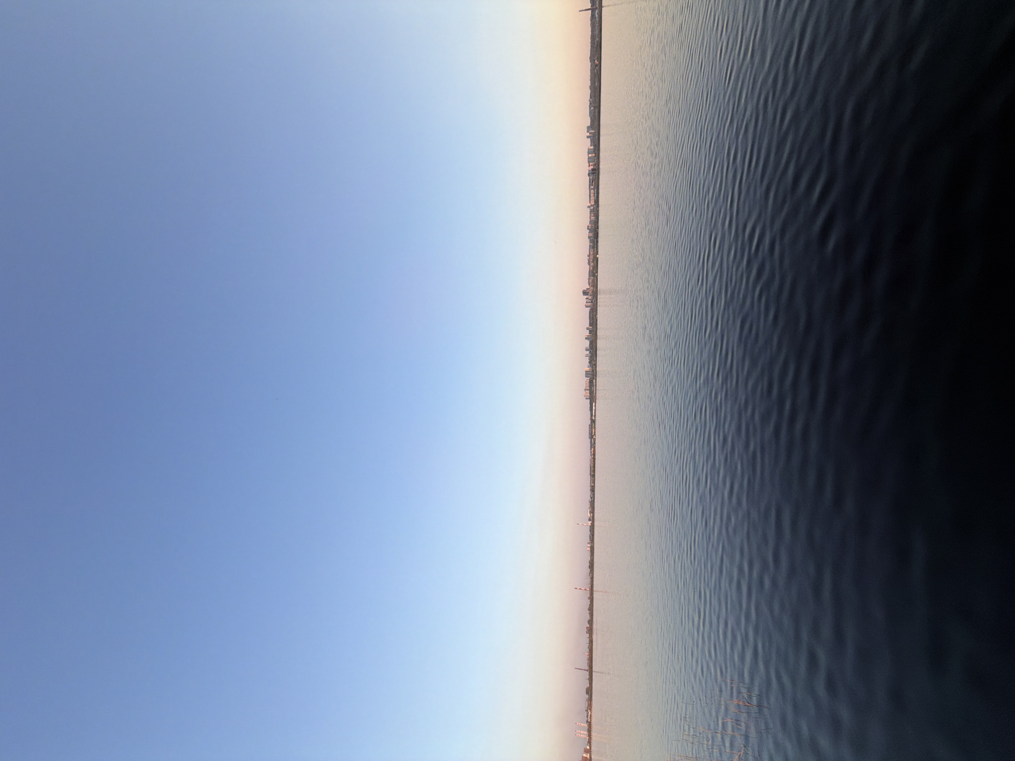 The image presents a view of a cityscape across a body of water. The sky is a clear gradient of blue, transitioning to lighter shades near the horizon. A city skyline is visible across the water, featuring a mix of high-rise buildings and industrial structures. The water surface is textured with small waves. The photograph is taken horizontally. There are no people visible. The location is most likely Izhevsk, Russia.