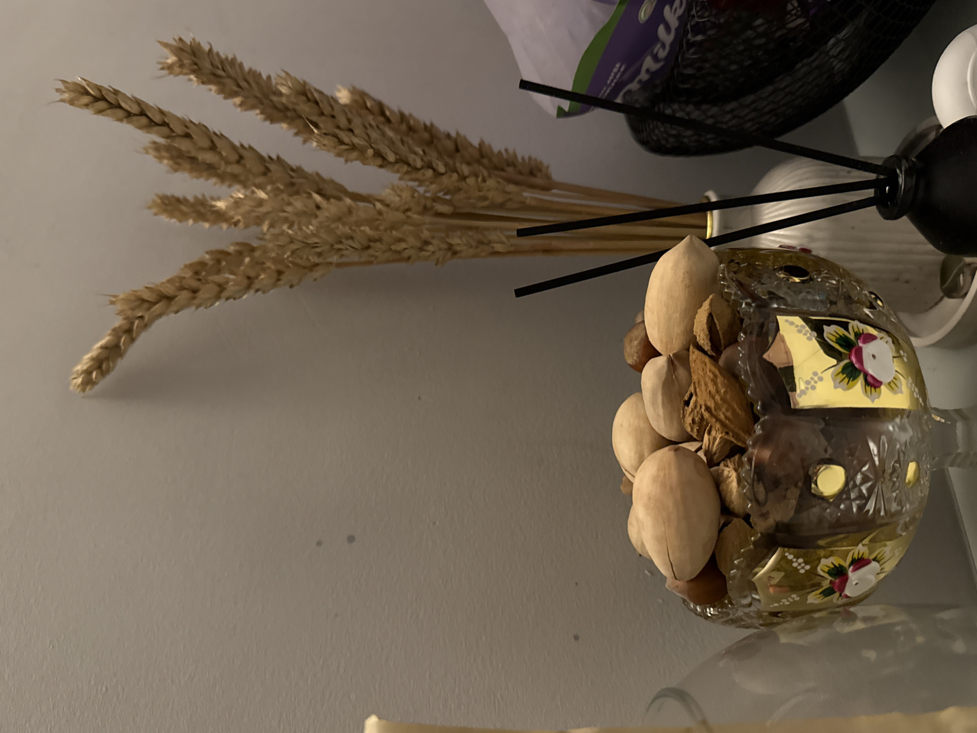 The image depicts a still life scene. A bundle of wheat stalks is positioned on the left side of the frame. Adjacent to the wheat, there's a decorative glass container filled with various nuts. The container is adorned with floral patterns. Behind these objects, elements of a household setting are visible, including what appears to be part of a shelf and various containers.