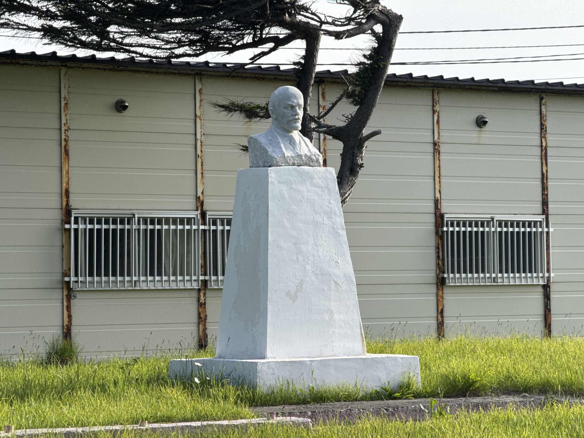 A bust of Vladimir Lenin is mounted on a white pedestal in front of a light-colored, corrugated metal building. The building has two windows with metal bars and what appear to be security cameras mounted on the exterior walls. A gnarled tree with dark foliage stands behind the bust, partially obscuring the building. The ground in front of the building is covered in lush green grass. The scene is set under an overcast sky.