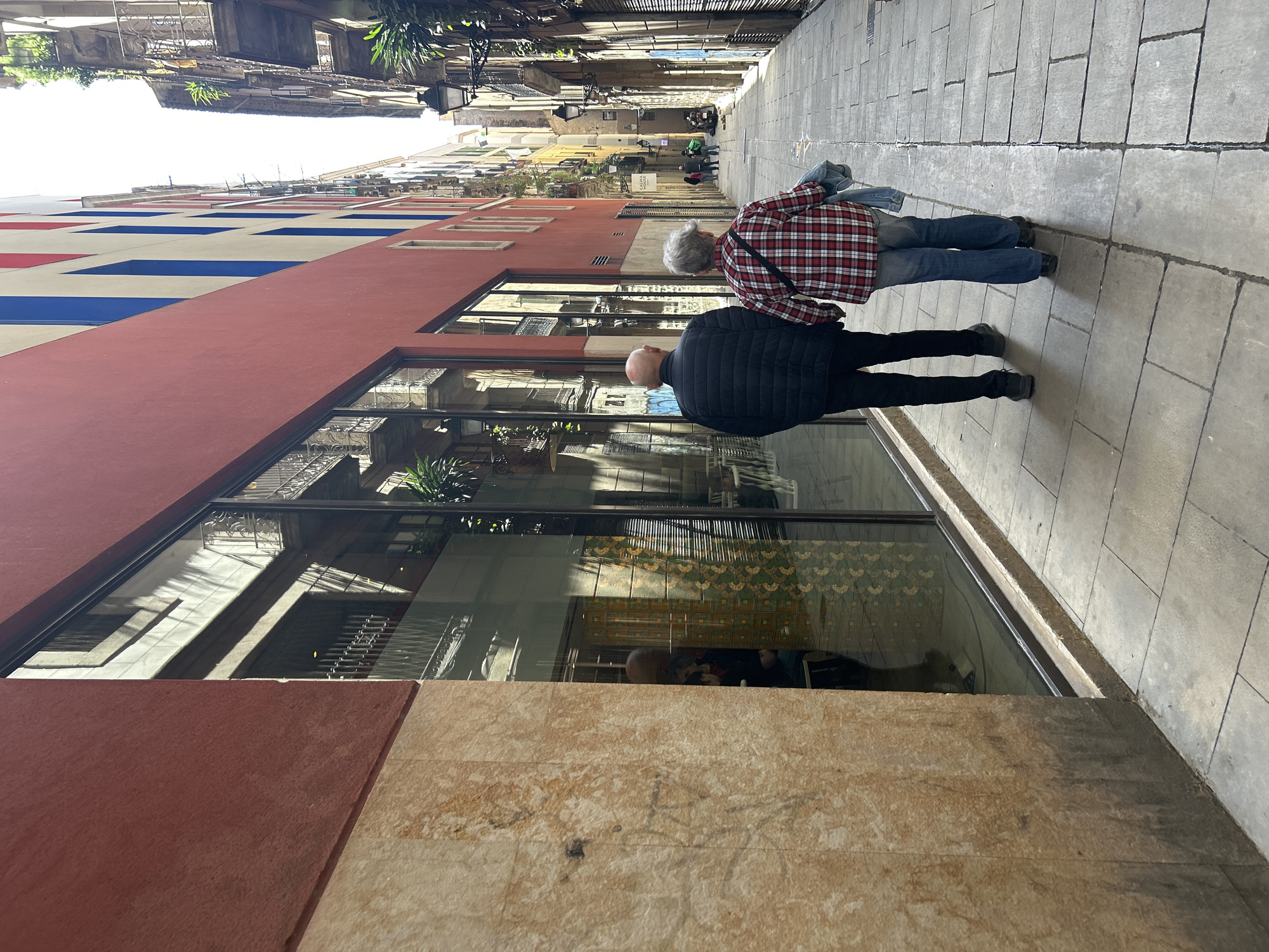 The image depicts a sunlit, narrow pedestrian street in a city, likely Barcelona, viewed from a slightly elevated perspective. Two individuals are walking away from the camera, heading down the street. The person in front is wearing a black puffer jacket and dark trousers, and appears bald. Behind them, another person is dressed in a red and white plaid shirt, blue jeans, and has short grey hair, with a denim jacket draped over their arm.
The street is paved with grey flagstones, showing distinct patterns and seams. On the left side, a prominent building features a large red lower section, with its upper facade decorated with bold red and blue rectangular patterns against a lighter background. This building has large ground-level shop windows, reflecting the architecture of the opposite side of the street, showing sunlight, shadow patterns, and a glimpse of what appears to be an ornate, patterned ceiling or interior detail within the reflection.
Further down the street, more traditional, taller buildings with balconies and shutters line the path, and other pedestrians are visible in the distance. The bright conditions indicate it is daytime, with clear sunlight casting distinct shadows. A sign hanging from a building further down the street prominently displays the words "CALMA MATEU".
