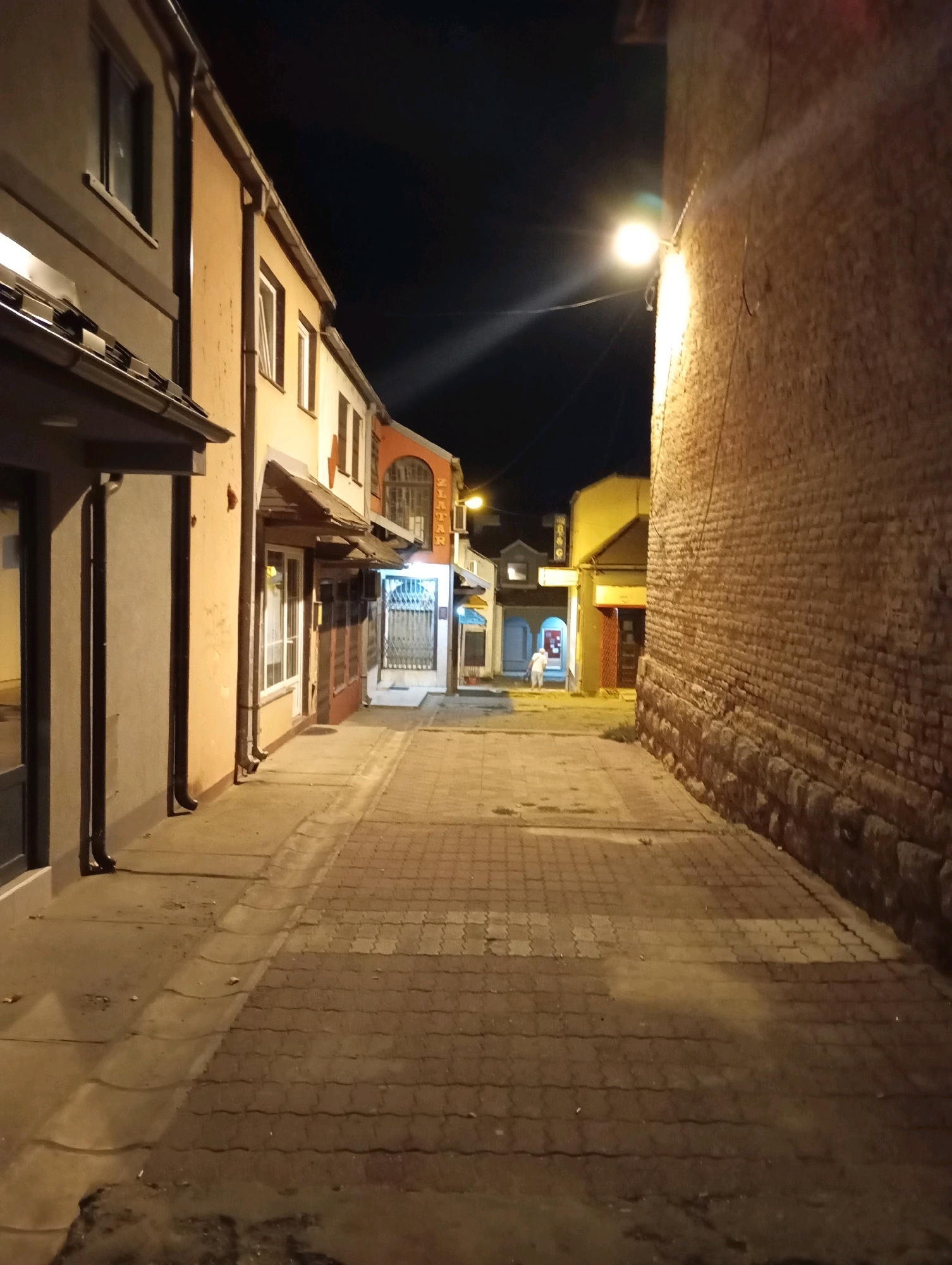 The image depicts a narrow, empty cobblestone alleyway at night in Topola Municipality, Serbia. The alley is flanked by buildings on both sides, with a tall brick wall dominating the right side of the frame. A street lamp on a wire illuminates the scene, casting long shadows. The buildings on the left appear to be commercial establishments with windows and awnings. Further down the alley, lights are visible from other buildings, suggesting more shops or residences. The overall impression is one of a quiet, possibly historic, urban environment after dark. There are no people clearly visible in the foreground, though a faint silhouette of two individuals can be seen in the distance. The ground is paved with interlocking stones and there are some fallen leaves and debris.