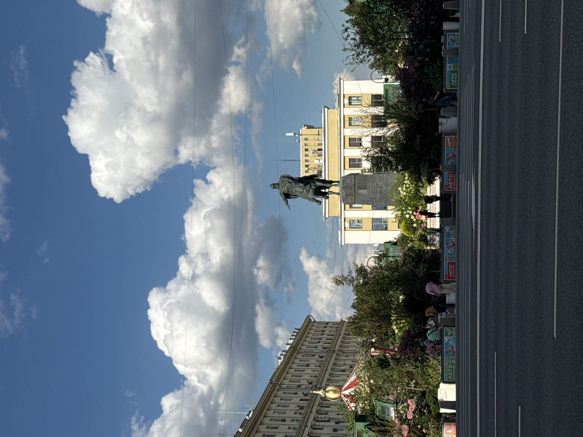 This is a vertical, medium-shot, eye-level photograph of a city street in Moscow, Russia, on a sunny day with a blue sky and large white clouds. In the foreground is a dark asphalt road with white lane markings. Behind the road is a raised sidewalk area with planters containing lush green foliage and colorful flowers, along with several people. To the left of the planters is a portion of a large, ornate building with a golden dome. In the center of the image, behind the planters, is a large bronze equestrian statue of a man in medieval armor. Behind the statue is a light yellow building with a flat roof and numerous windows. Further in the background, on the left side of the frame, is another long, pale building with many windows, and overhead power lines are visible against the sky. The atmosphere is bright and vibrant, with a mix of urban architecture and greenery.