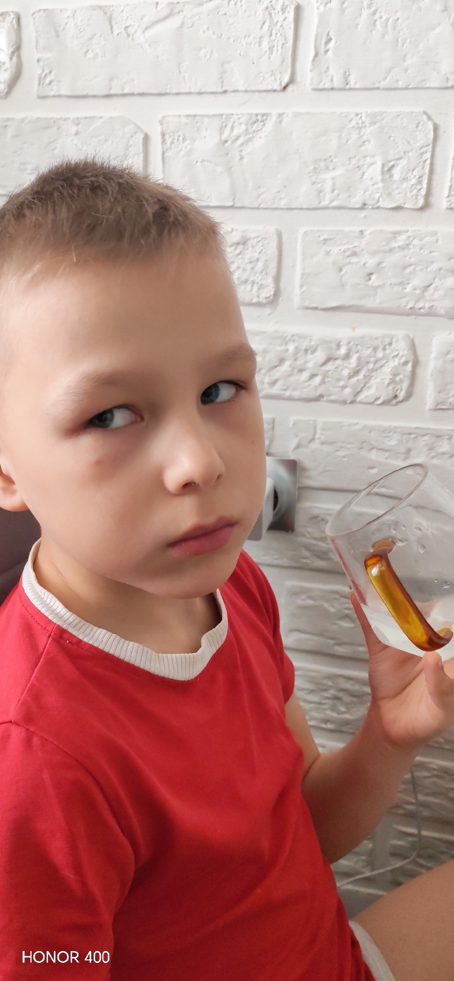A young boy with light brown hair and blue eyes is holding a clear glass with an amber-colored handle. He is wearing a red t-shirt with a white trim. He is looking directly at the camera with a slight expression. Behind him is a white brick wall with an electrical outlet. The text "HONOR 400" is at the bottom left corner. The location is likely indoors. It is impossible to verify the city of Noviy, Russia with the information given.